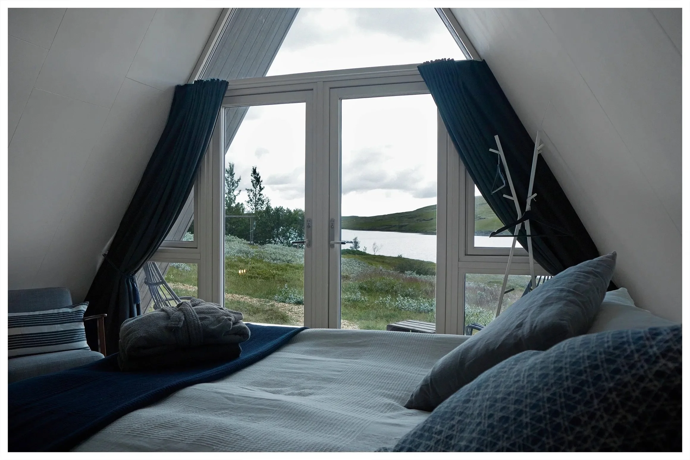 A cozy bedroom with a large window overlooking a lake and green landscape, with dark curtains on either side, a bed with pillows and a folded towel, and a drying rack in the corner.