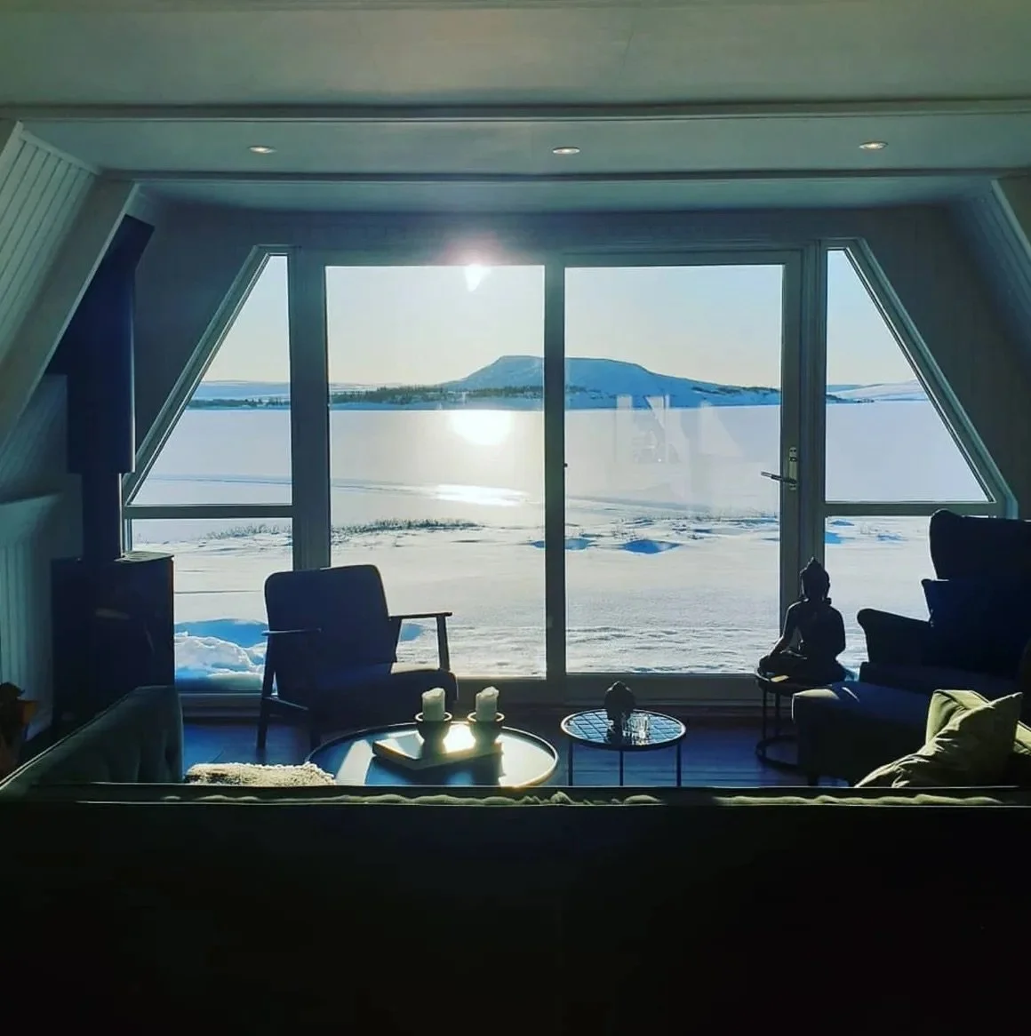 Living room with large window and view of snowy landscape, mountains, and water outside. Furniture includes chairs, a sofa, and small tables with candles and decorative items.