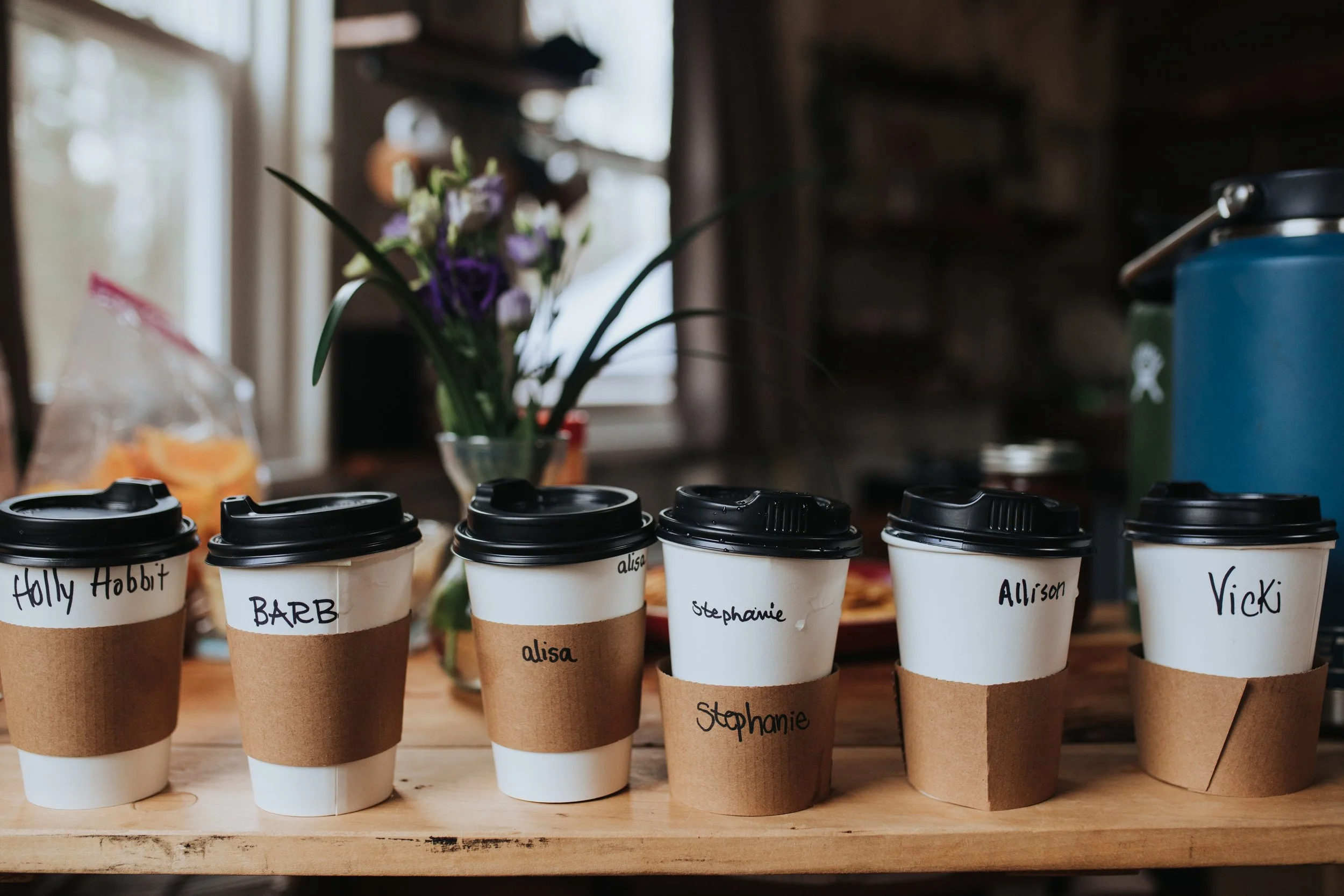 Multiple disposable coffee cups with black lids and handwritten names on the sleeves, arranging from left to right: Holly Hobbit, Barb, alisa, Stephanie, Allison, Vicki, in front of a kitchen window with a vase of purple and white flowers and various kitchen objects and furniture.