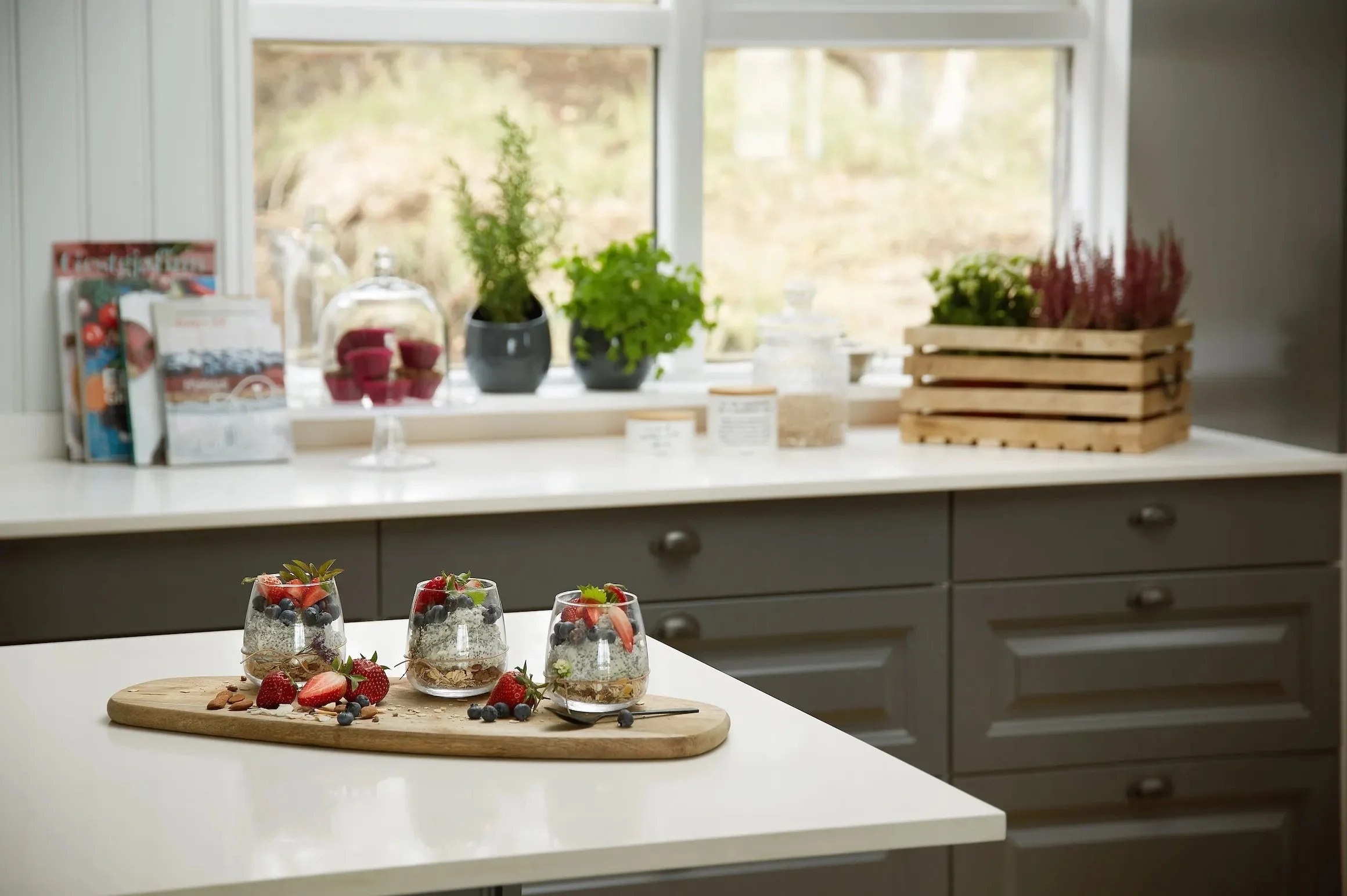 Three dessert parfaits in glasses topped with strawberries, blueberries, and chocolate shavings, arranged on a wooden board with additional berries around.