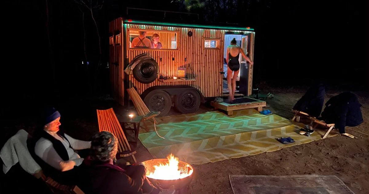 People gathered around a campfire near a mobile tiny house with a woman standing on the steps. The tiny house has a wood exterior with colorful LED lights and a window with a person inside. The scene appears to be at night.