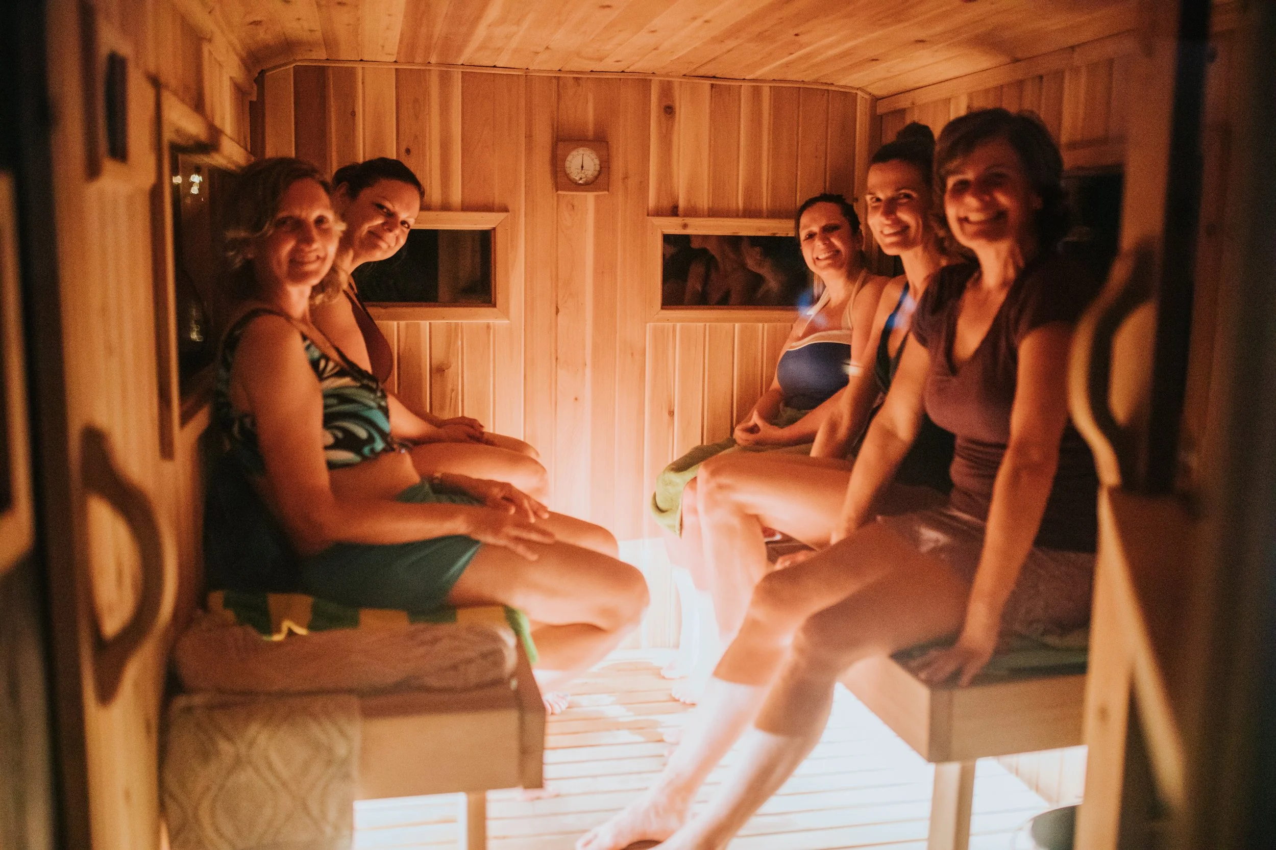 Five women sitting inside a wooden sauna room, smiling at the camera, with a clock on the wall behind them.