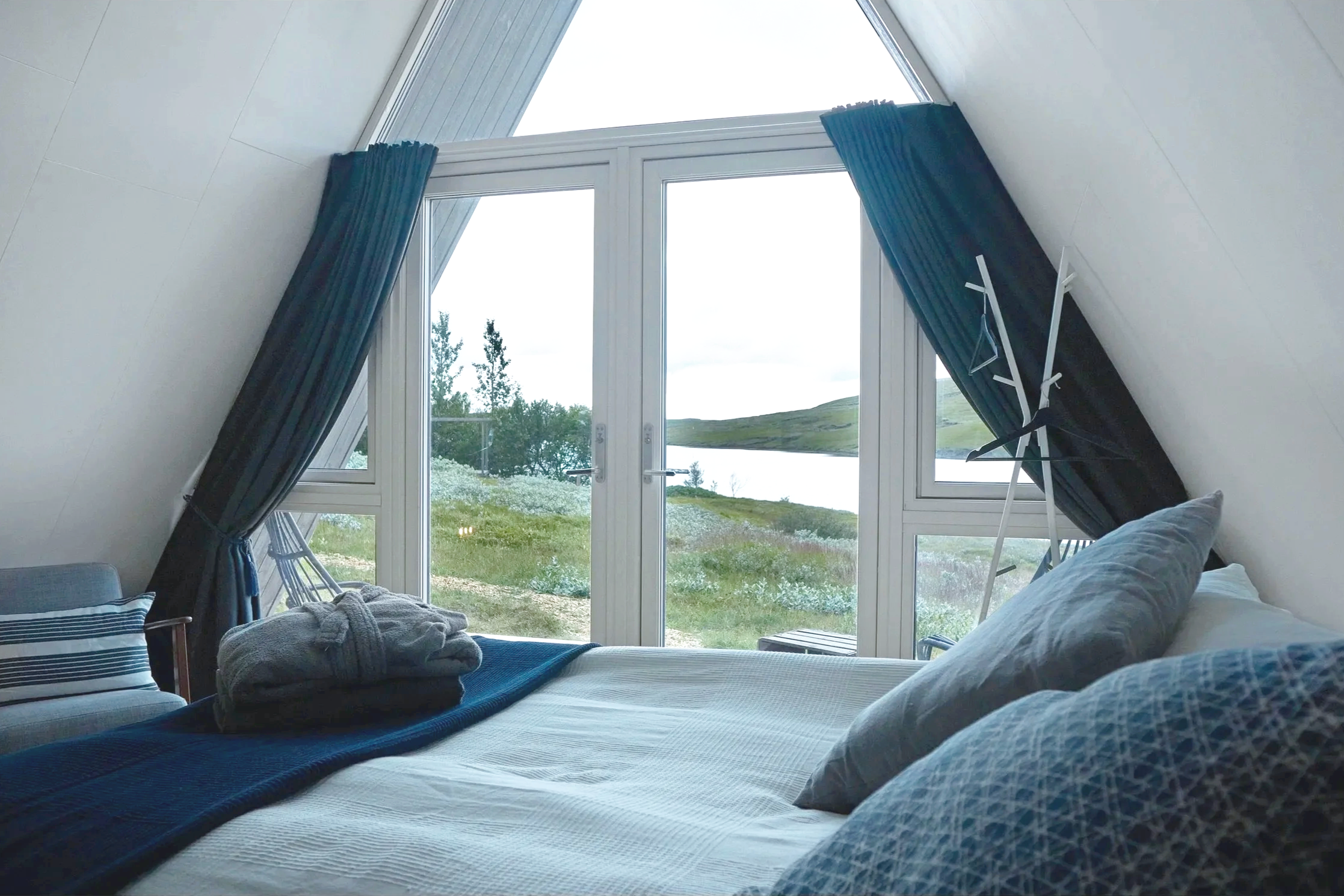 A bedroom with large glass doors overlooking a scenic outdoor landscape with grass, trees, and a body of water, under an angled roof with white walls and blue curtains.
