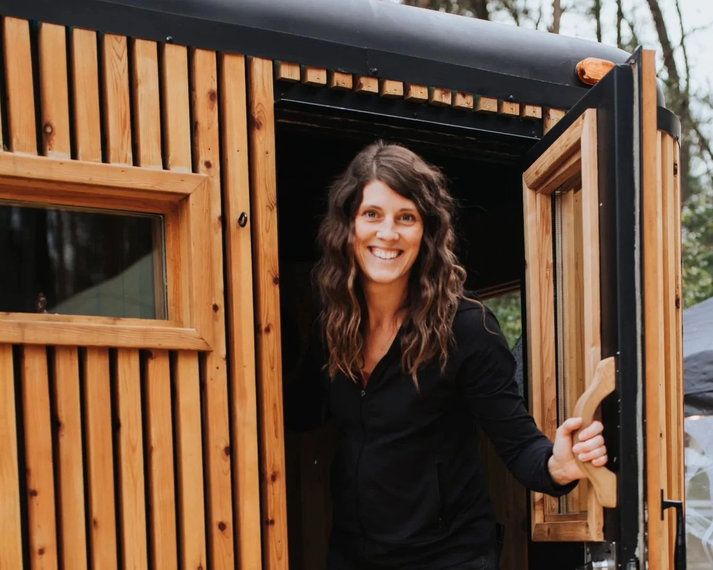 Woman with curly brown hair smiling as she leans out of a wooden door of a tiny house.