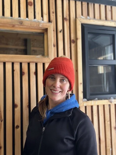 Woman smiling, wearing a red knit beanie and black jacket indoors with wooden walls and two windows.