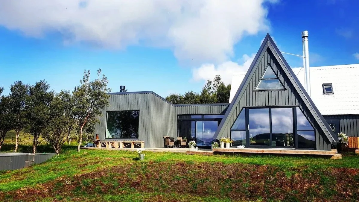 Modern house with a steep A-frame roof, large glass windows, and an outdoor deck, set in a green landscape with trees and a partly cloudy sky.