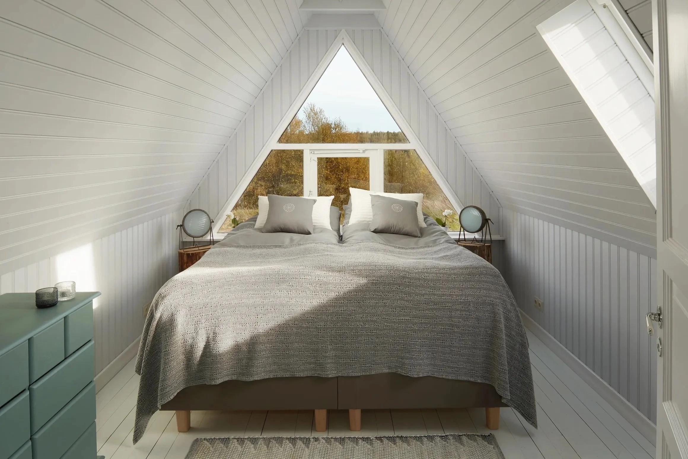 A bedroom with a triangular window behind a bed in an attic, decorated in neutral tones, with two bedside tables and lamps.