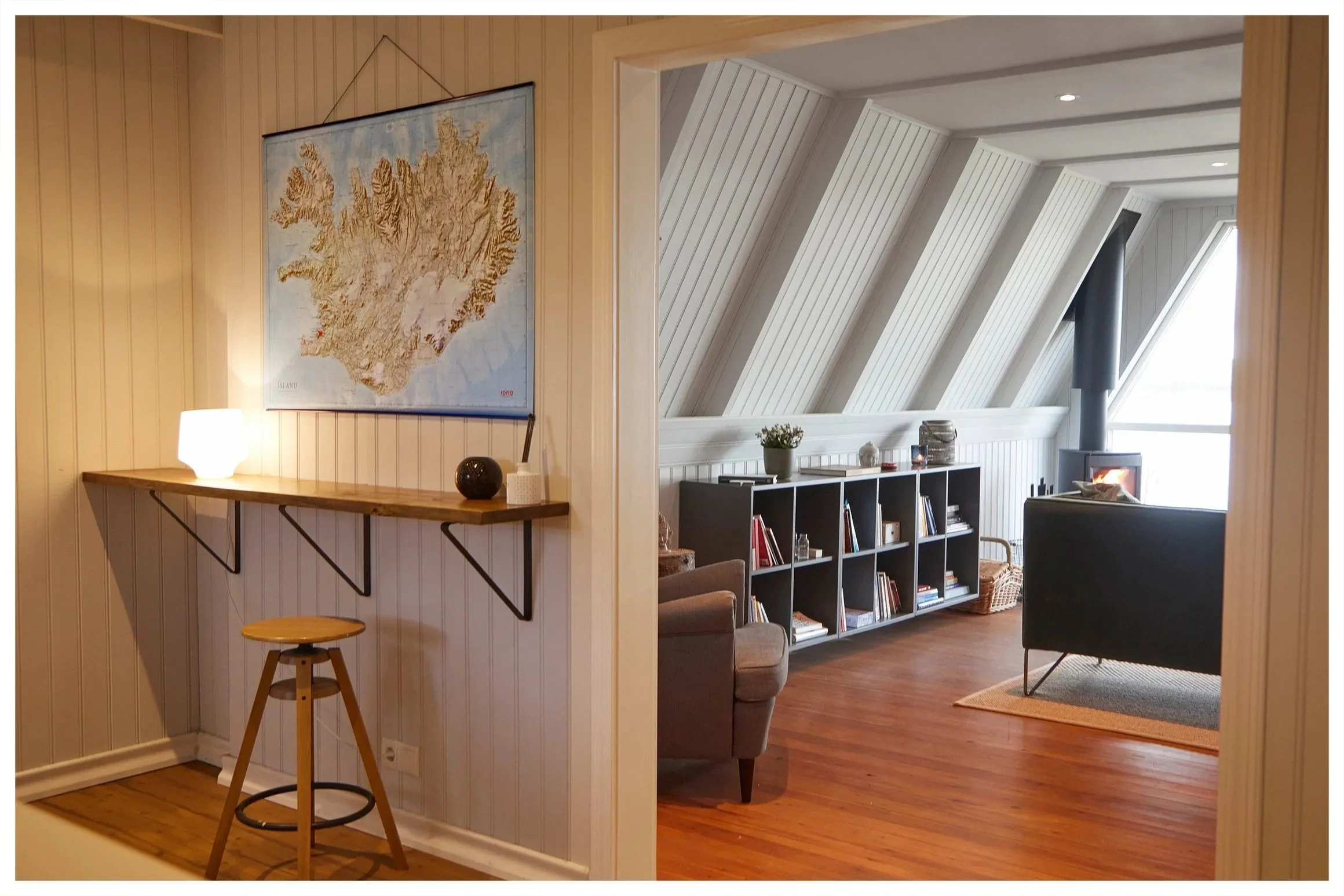 Interior view of a living room with a black bookshelf, gray armchair, wood floor, and fireplace, seen through a doorway from a small corner with a wooden stool and mounted map of Iceland.