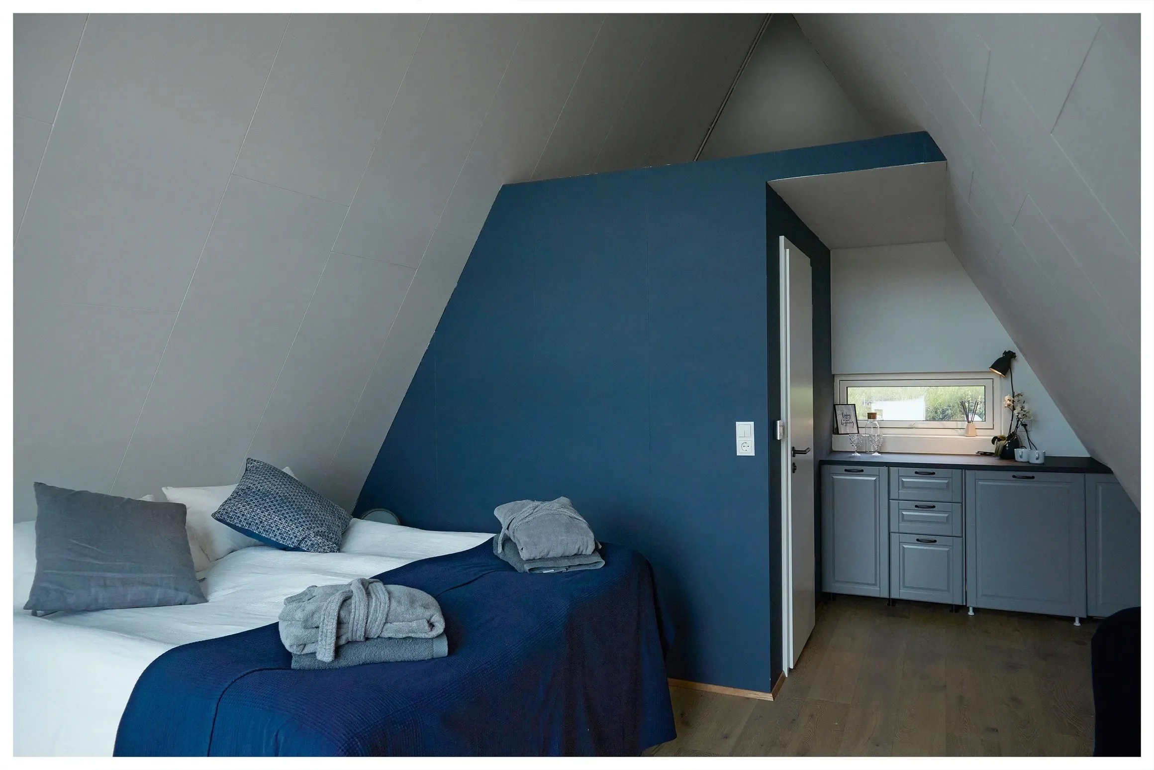 A bedroom with sloped ceiling walls. The bed has white sheets, gray and blue pillows, and navy bedding. Towels are folded on the bed. The room has a built-in gray cabinet under a small rectangular window with decorative items on the windowsill.