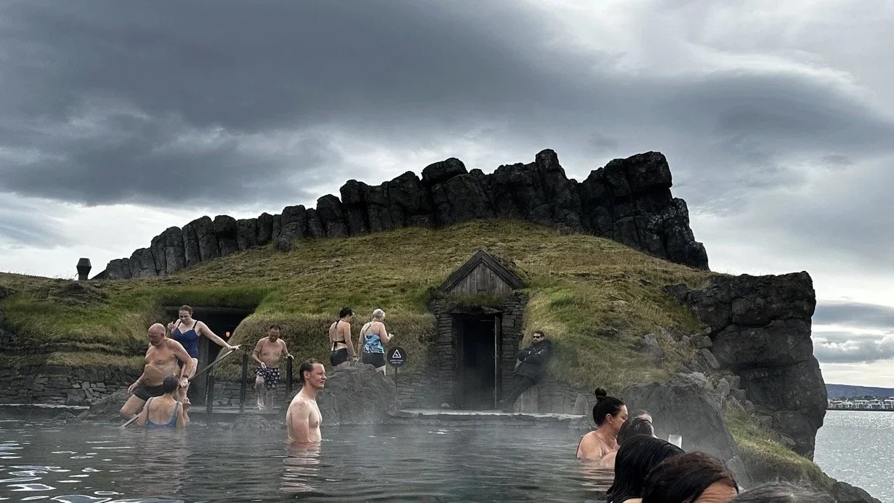 People bathing in a hot spring pool with a rocky hillside and cloudy sky in the background.