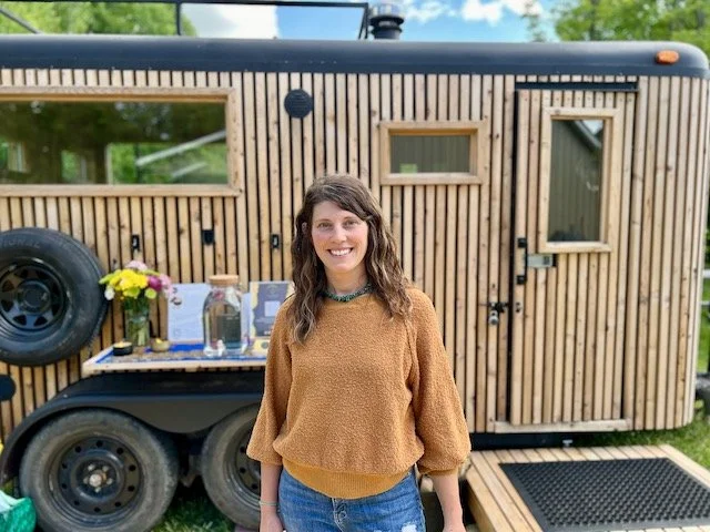 Woman standing outside in front of a wooden tiny house, smiling, with a table holding flowers and water bottles behind her.