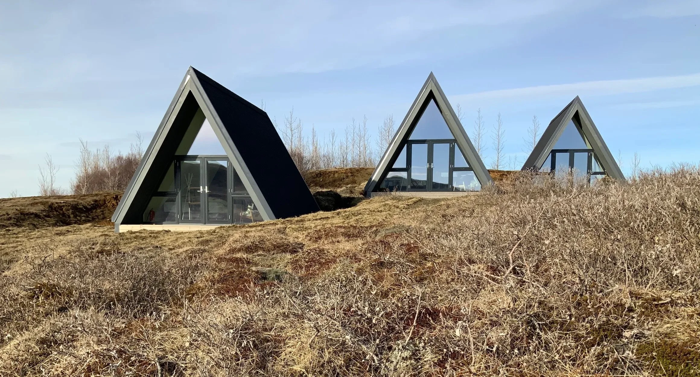 Three modern A-frame glass houses on a grassy hillside with dry shrubs, under a blue sky with light clouds.