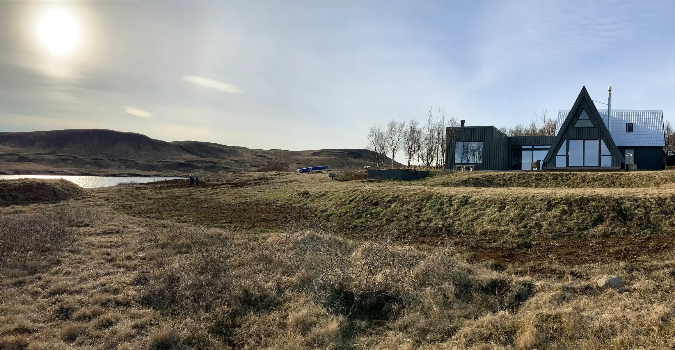 A modern black house with large glass windows and a steep roof, situated on a grassy hillside with leafless trees in the background and rolling hills under a partly cloudy sky.