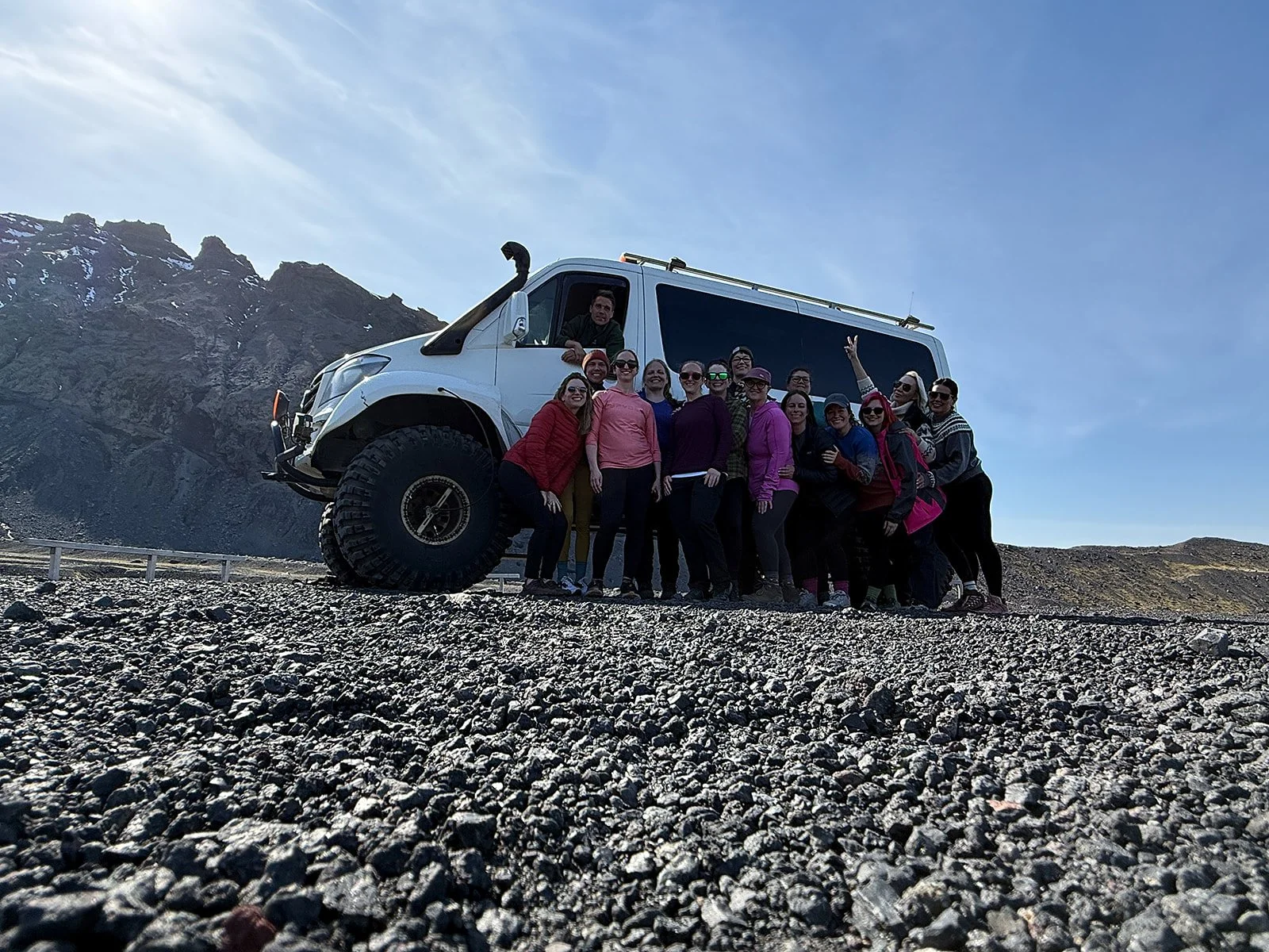 Group of people posing in front of a large white off-road vehicle on a rocky landscape with mountains in the background.