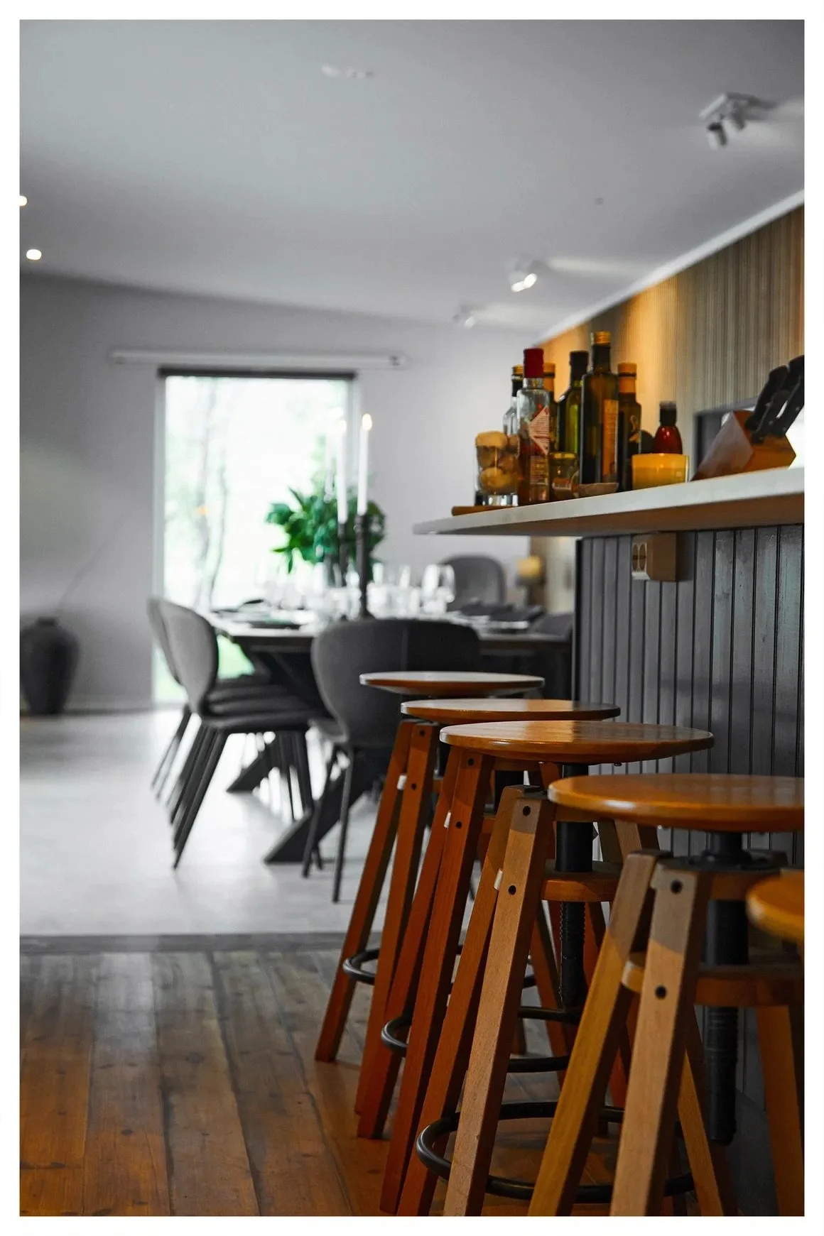 The interior of a modern restaurant or cafe with wooden bar stools along a counter, dark wooden chairs around a dining table, and a window in the background with greenery outside.