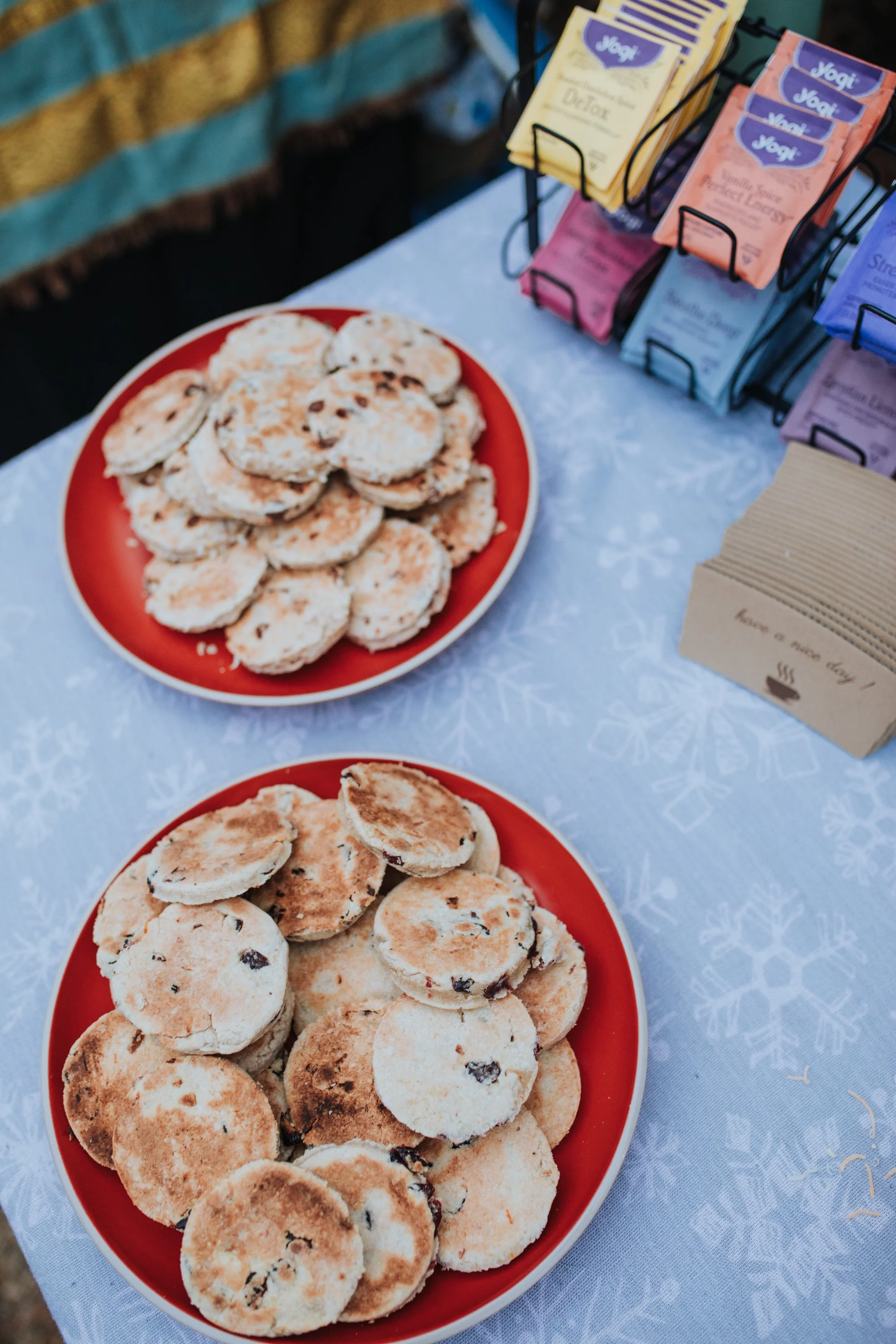 Two red plates filled with assorted cookies on a table covered with a white tablecloth, with a metal rack displaying colorful sugar packet labels in the background.
