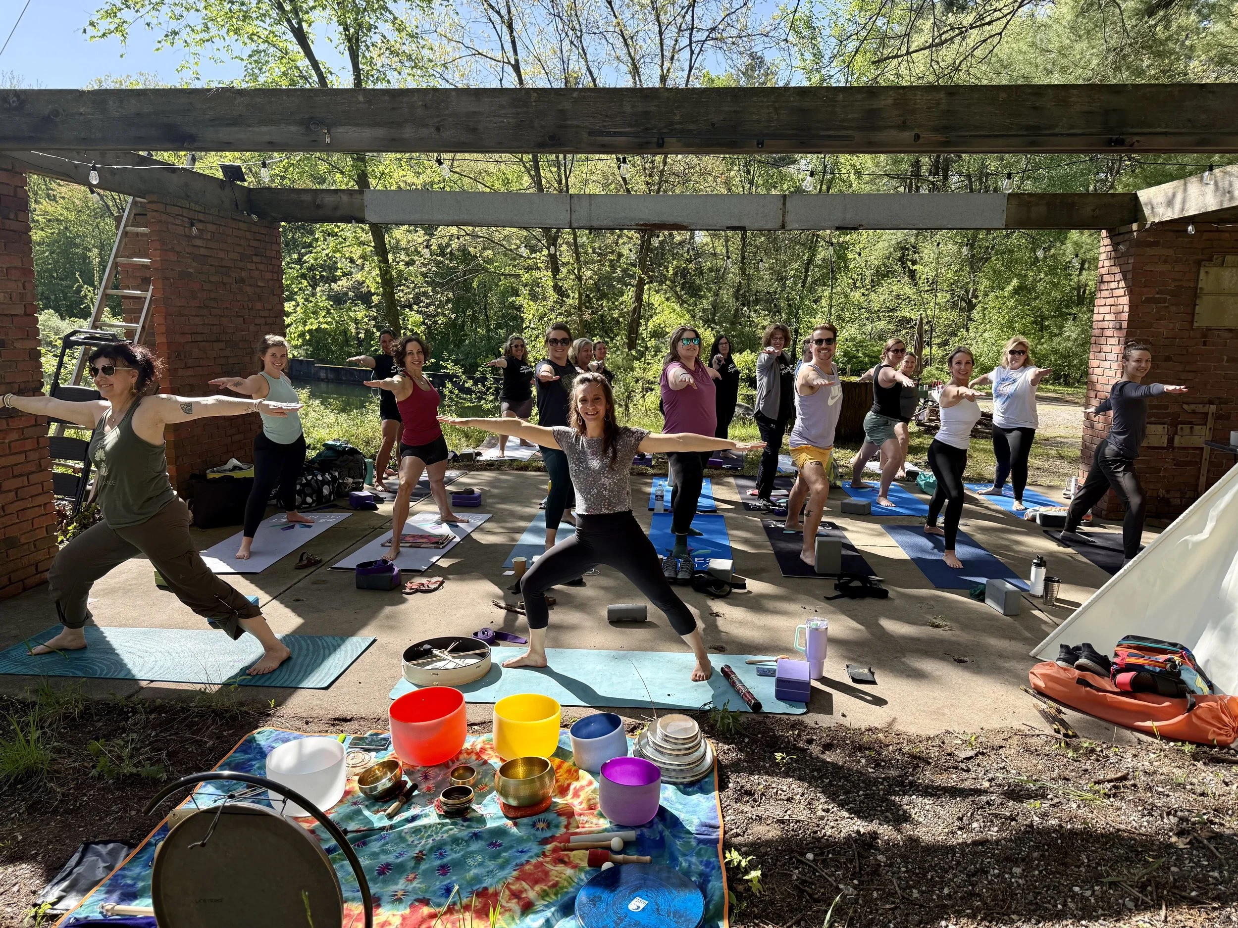 A group of people participating in an outdoor yoga class under a wooden pavilion in a forested area on a sunny day. They are standing on yoga mats with arms extended forward.