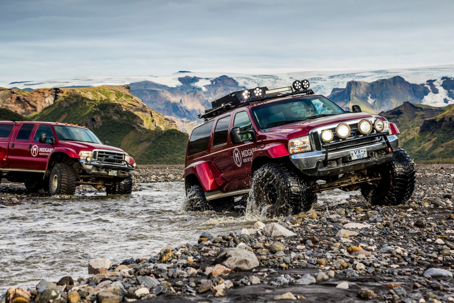 Two red off-road vehicles crossing a rocky river with mountainous landscape in the background.