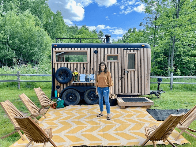 A woman standing outside a tiny wooden home with a yellow patterned outdoor rug, surrounded by trees and a wooden fence, on a sunny day.