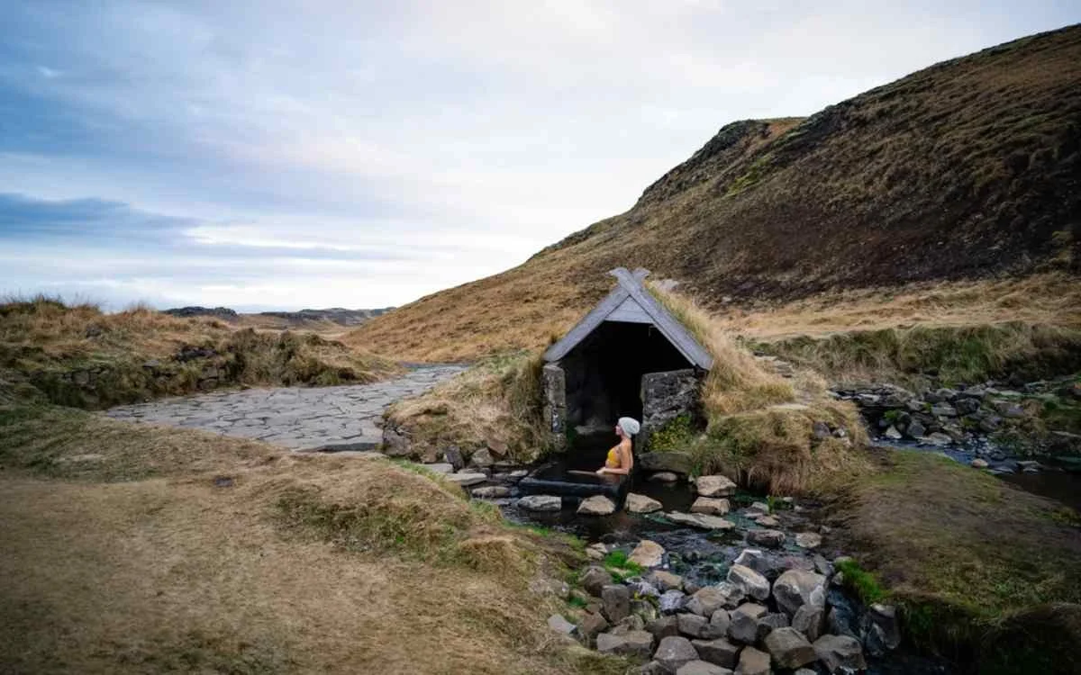 A person in a bathing cap sitting in a small, outdoor mineral hot spring near a stone building in a grassy landscape with hills and a cloudy sky.