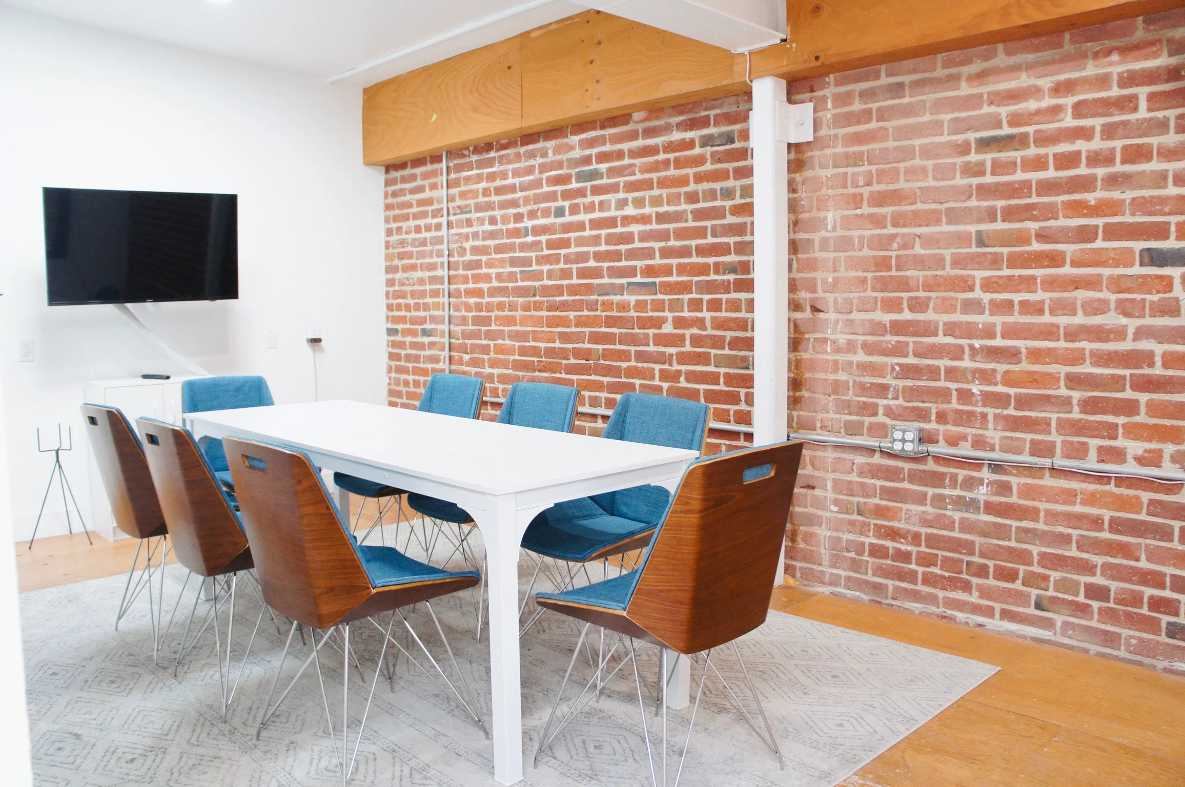 A modern conference room with a white table, six blue upholstered chairs, an exposed brick wall, a mounted flat-screen TV, and a white cabinet.