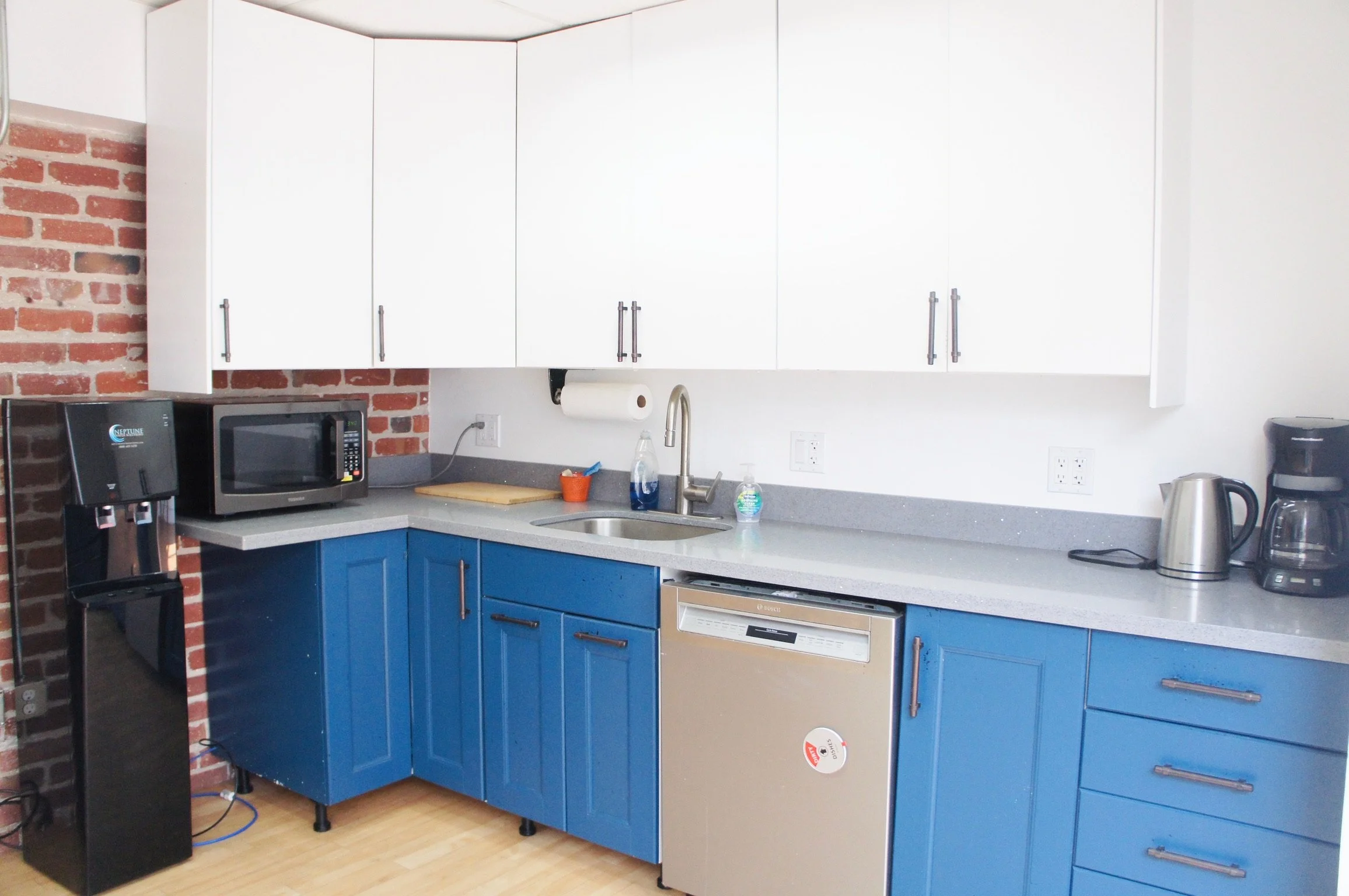 Kitchen with white upper cabinets, blue lower cabinets, and brick wall on the left. Contains a coffee machine, microwave, water cooler, electric kettle, and coffee maker on the countertop. There is a sink with a faucet, a paper towel roll, hand soap, and a bottle of water. Dishwasher is integrated into the blue cabinets.