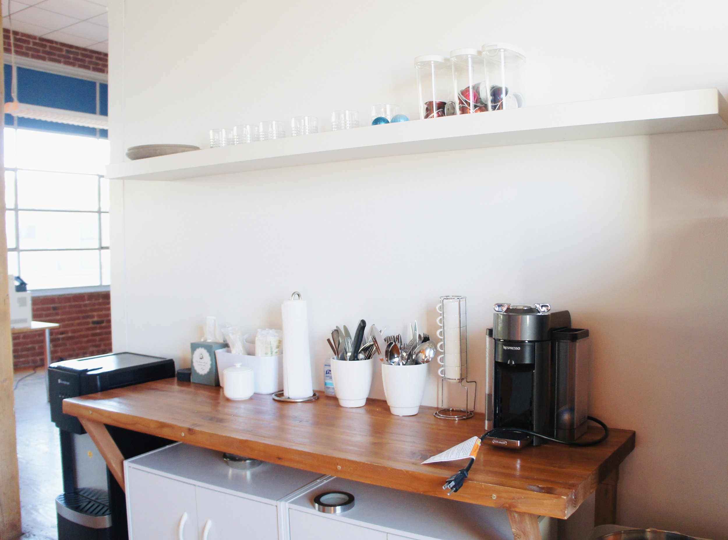 Coffee station with a Keurig machine, cups, utensils, and a countertop on a white wall, in an office or kitchen space.