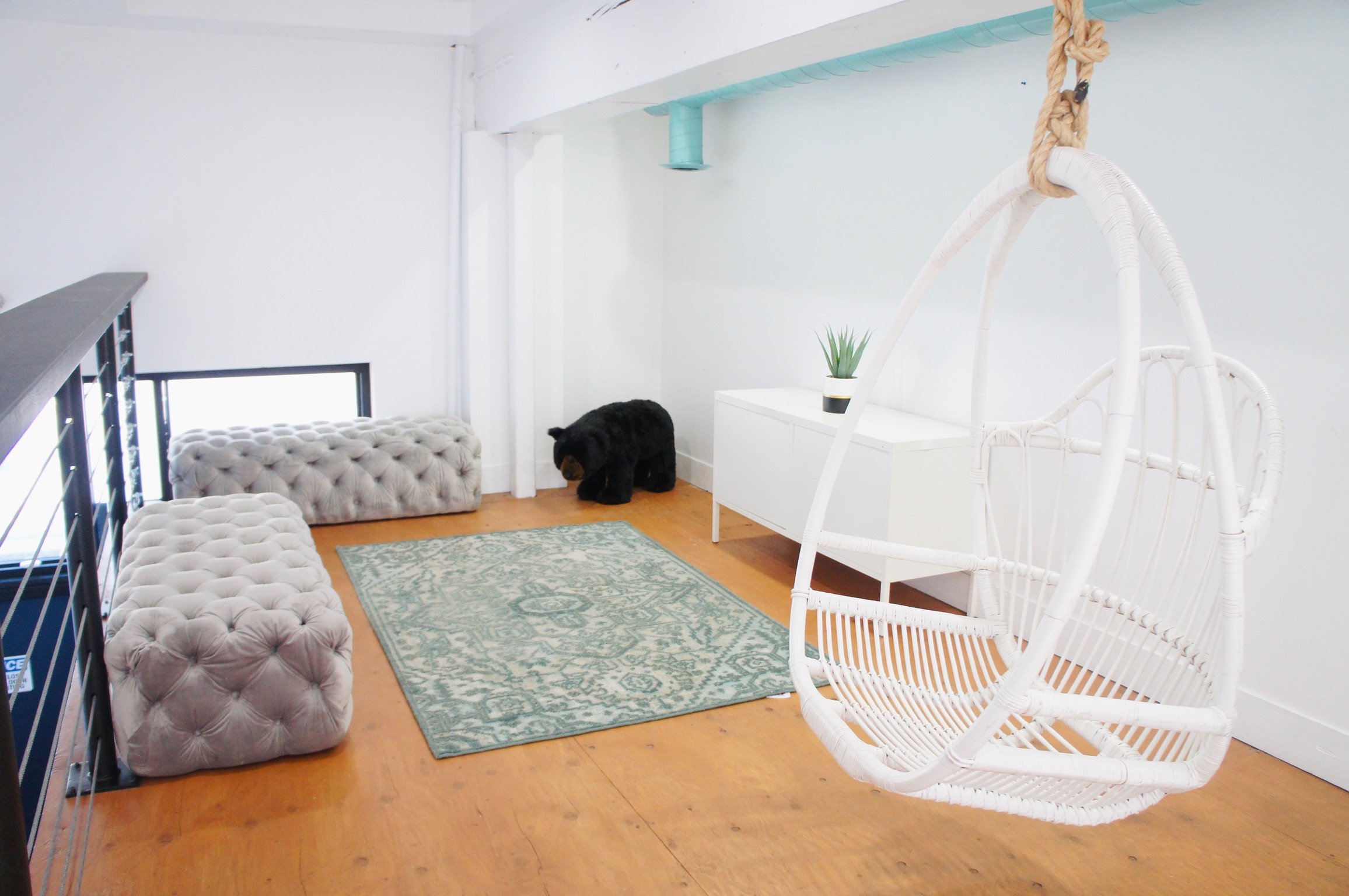 Interior of a modern living space with a white hanging wicker chair, tufted ottomans, an area rug, a black plush bear, a white side table with a plant, and light wood flooring.