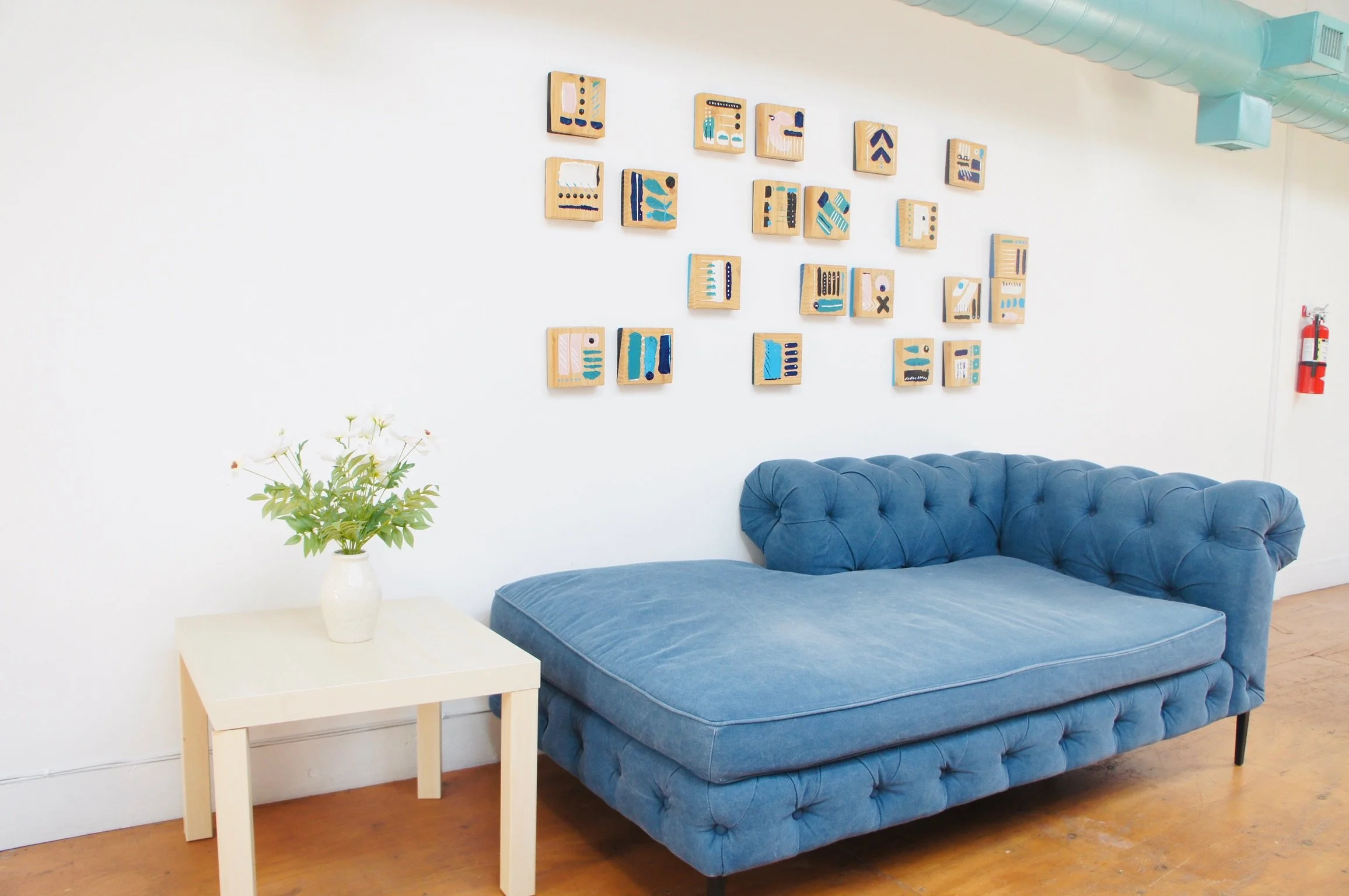 A cozy living space with a white wall adorned with small, abstract art pieces arranged in a pattern. A blue velvet tufted couch sits against the wall, next to a small white side table with a white vase holding green leafy plants. A fire extinguisher is mounted on the far right wall, and an air duct runs along the ceiling.