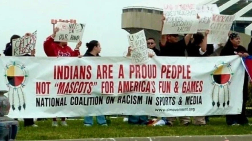 Group of people holding signs and a large banner at a protest or rally. The banner reads 'Indians are a proud people not 'mascots' for America's fun & games' with references to a national coalition on racism in sports and media.