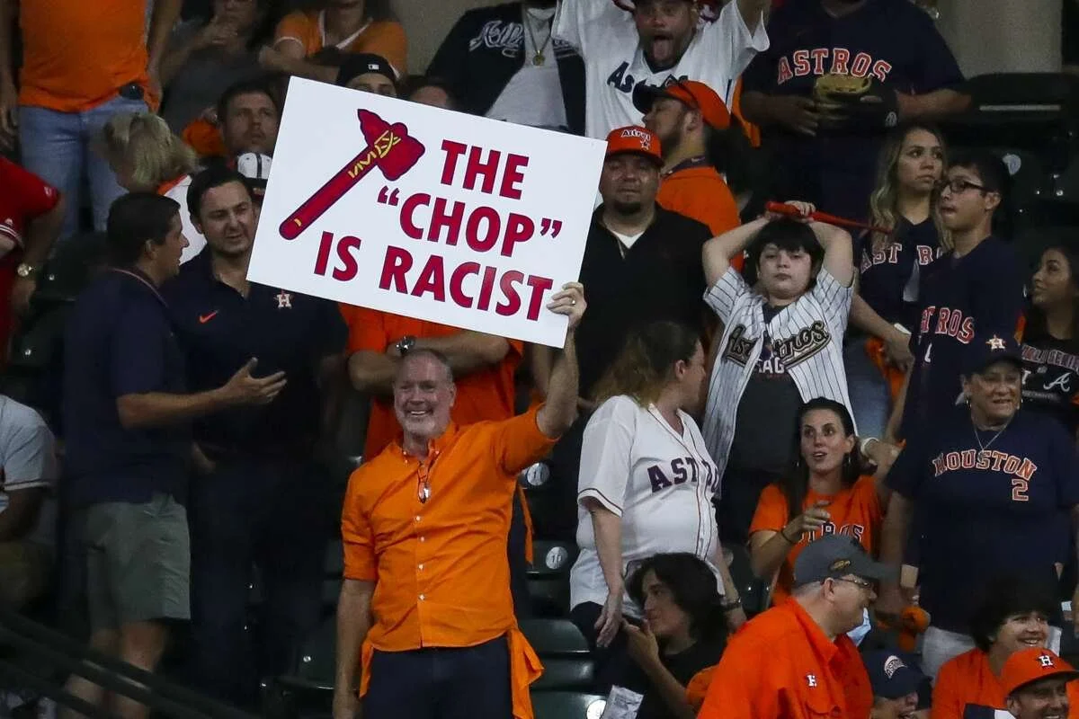 A man holding a sign that says, 'The Chop is Racialist,' written in red and white, with an axe illustration, at a baseball game with fans wearing Atlanta Braves apparel.