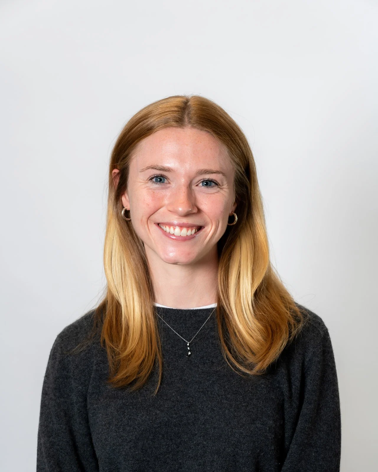 Portrait of a young woman with red hair, blue eyes, wearing a dark sweater, earrings, and a necklace, smiling against a plain white background.