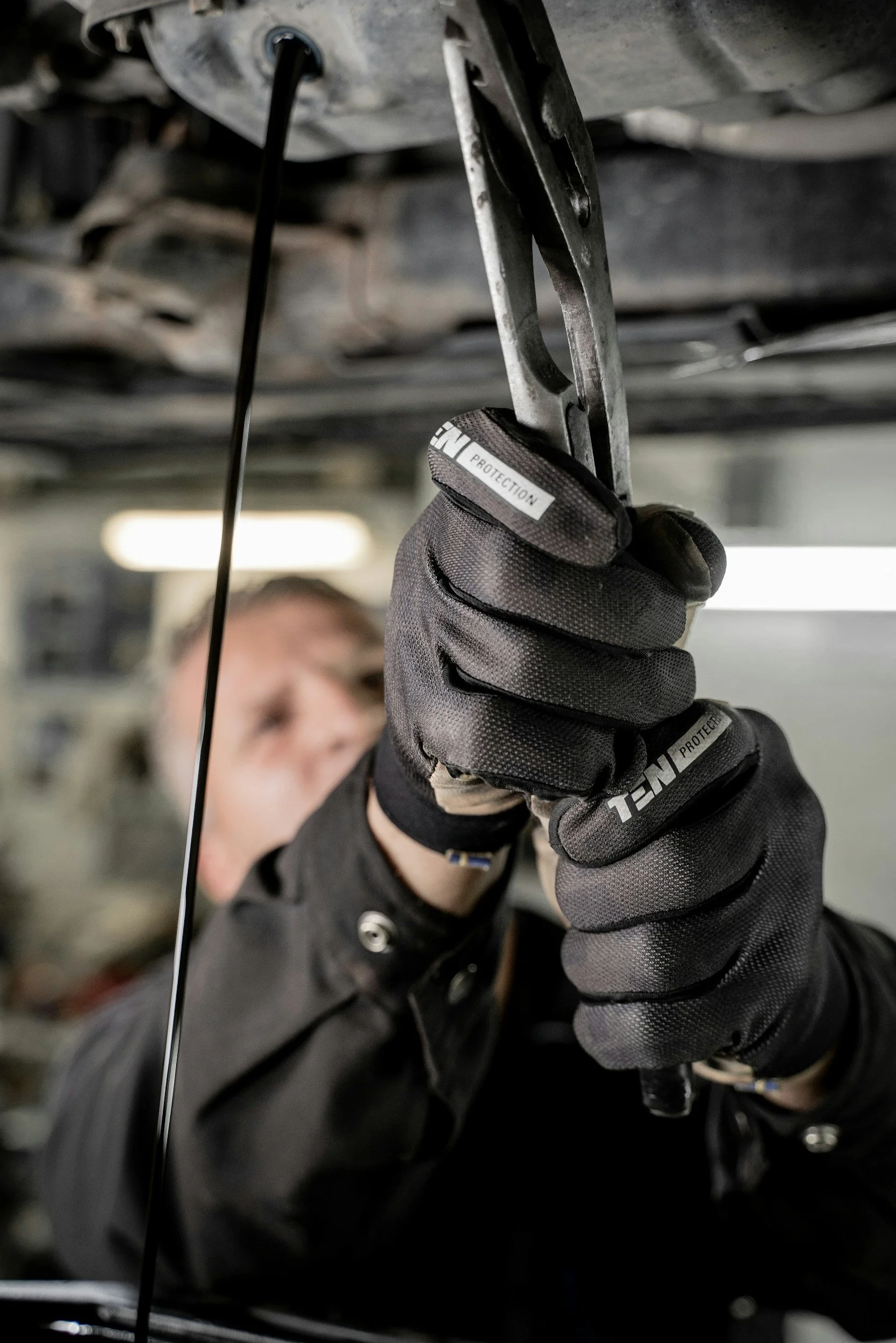 A person wearing black gloves working on a car engine, using a tool under the vehicle's chassis.