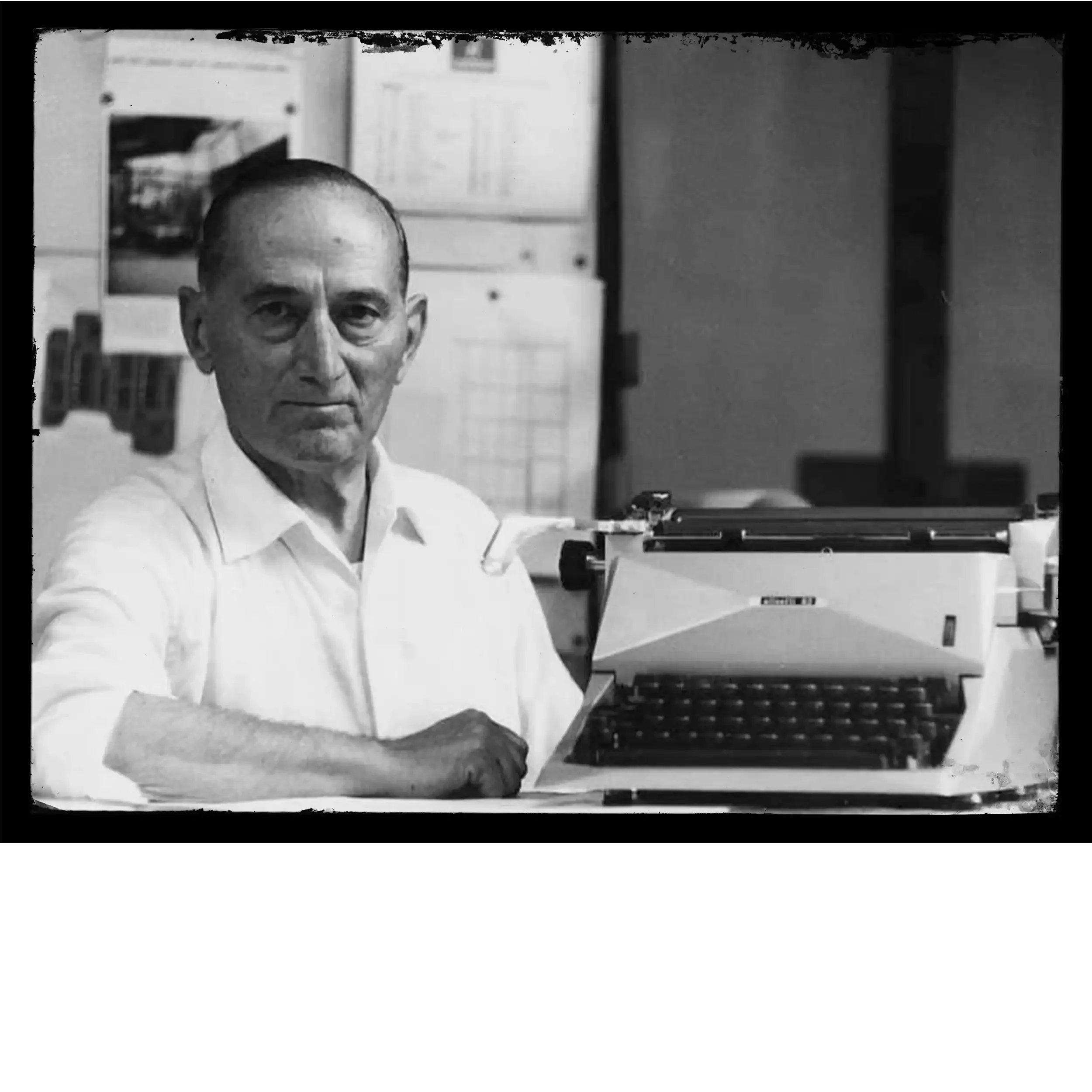 Black and white photo of a man with a serious expression, wearing a white shirt, sitting at a desk with a typewriter, with shelves and papers in the background.