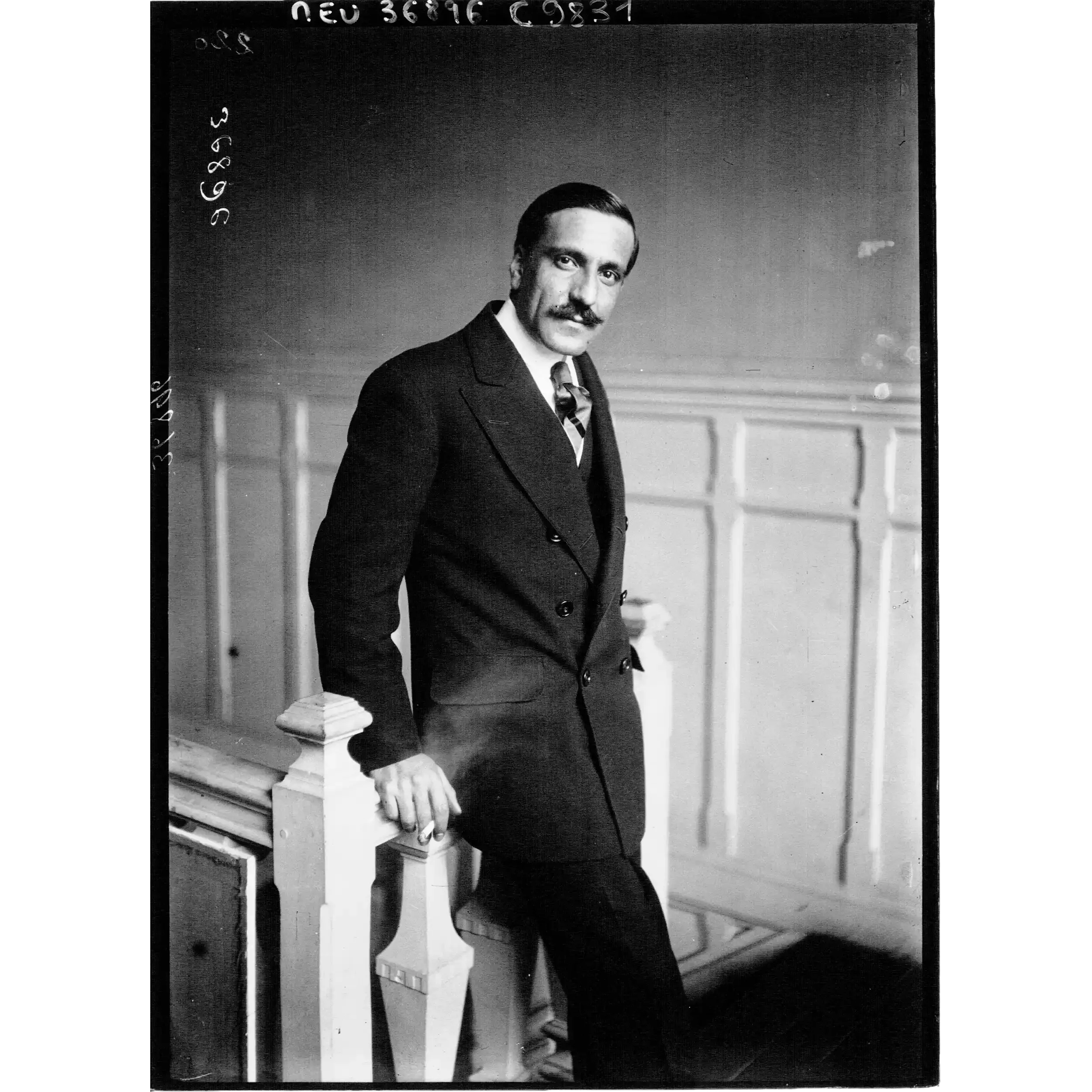 A black and white photograph of a man with a mustache and slicked-back hair, wearing a dark suit, standing next to a drawer or cabinet with multiple compartments. The background appears to be a plain interior setting. There is handwritten text upside down at the top of the image and on the left side.