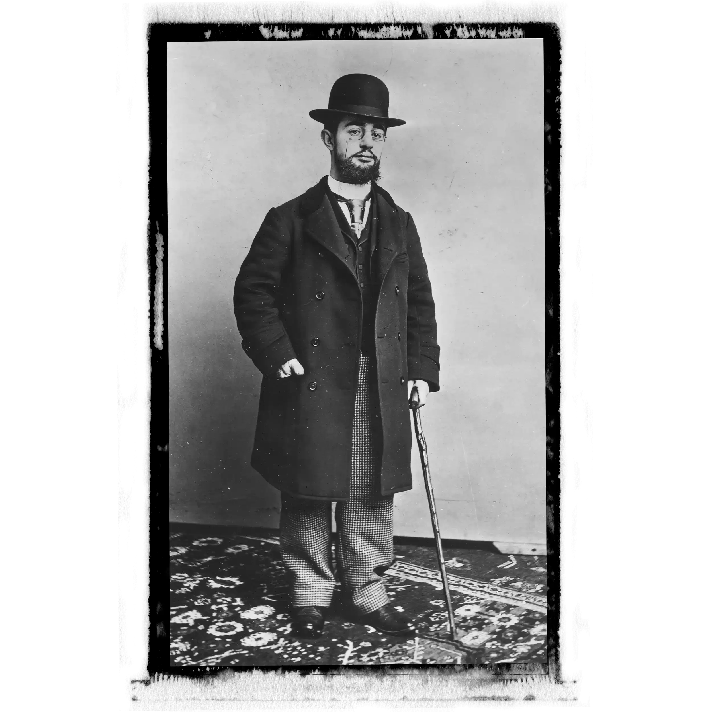 A black-and-white photograph of a man with a beard and glasses, wearing a bowler hat and a long coat. He is holding a walking stick and standing on a patterned rug.