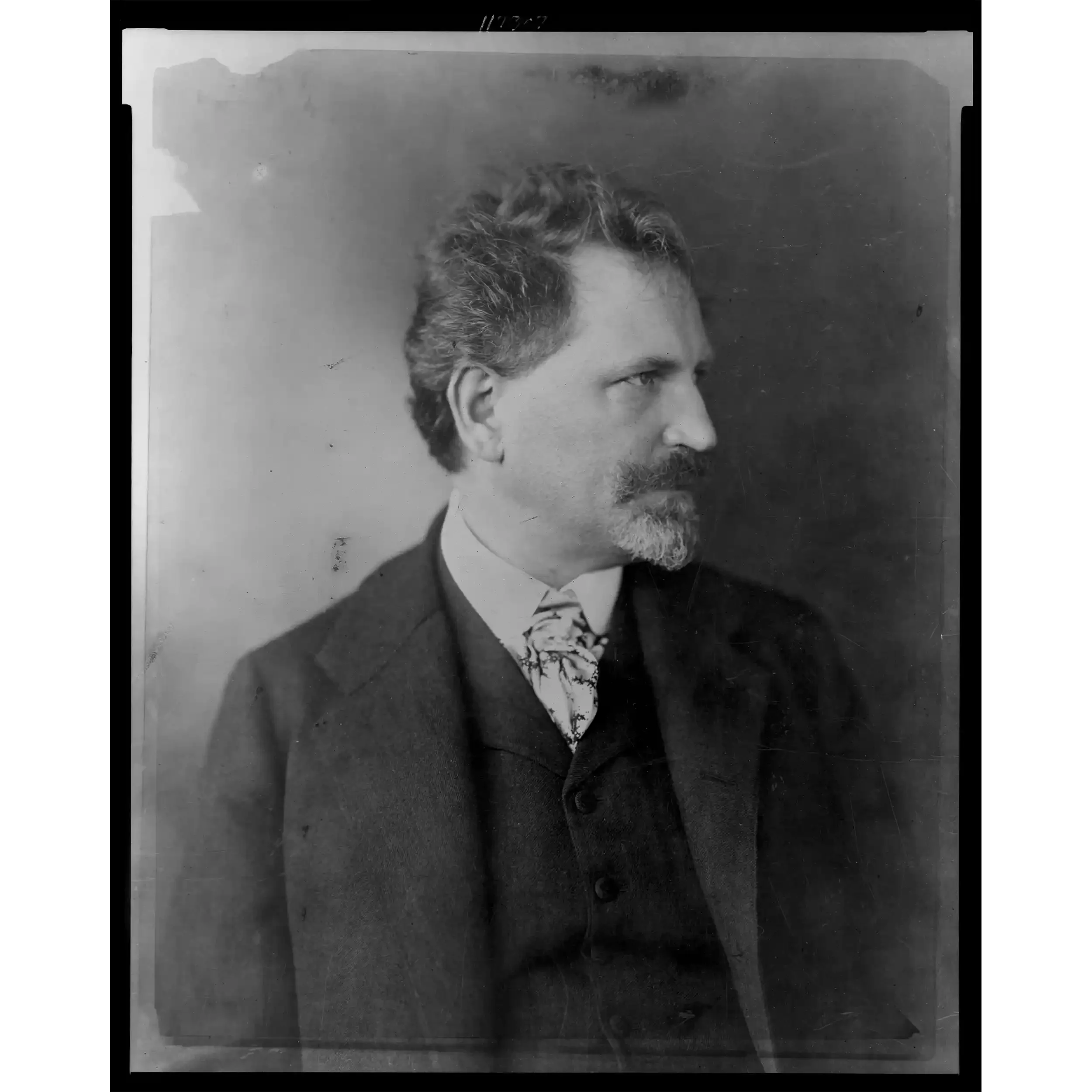 Black and white photograph of a man's profile with curly hair, a beard, and formal attire including a jacket and a patterned tie, looking to the right.