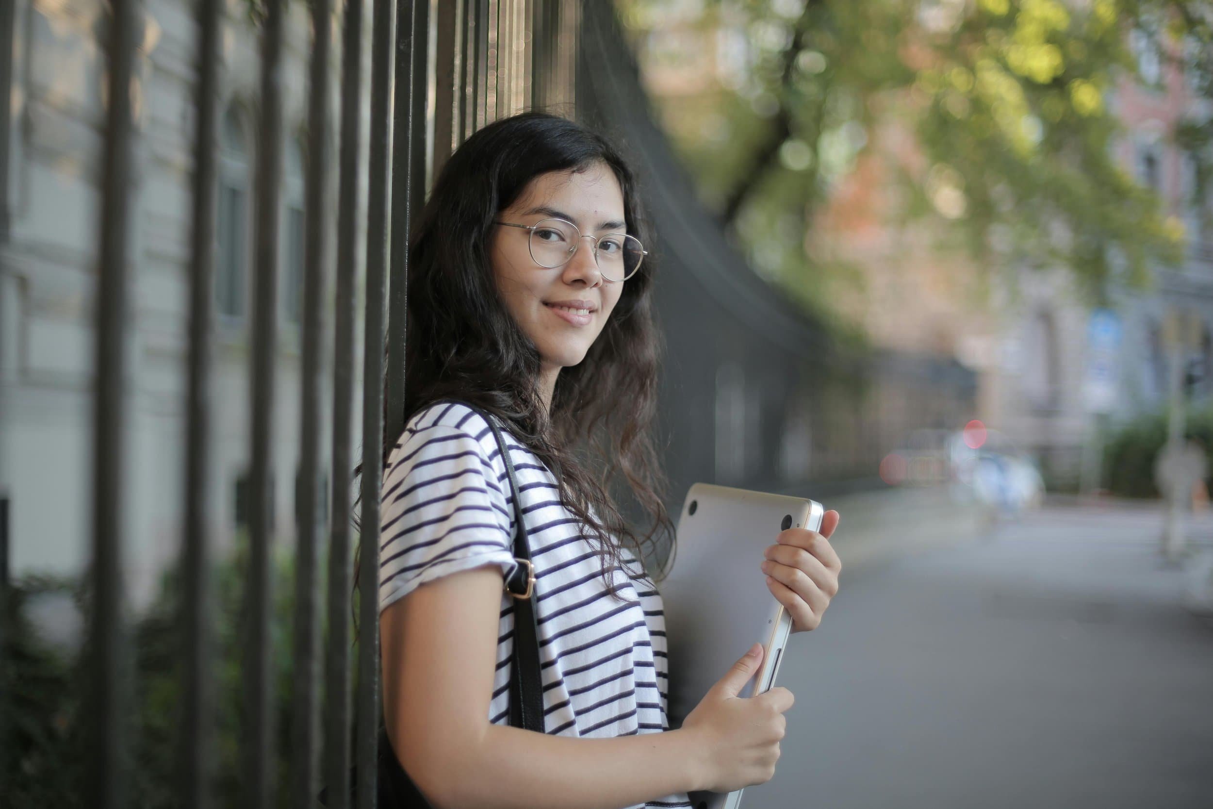 Woman smiling holding laptop leaning on fence in city