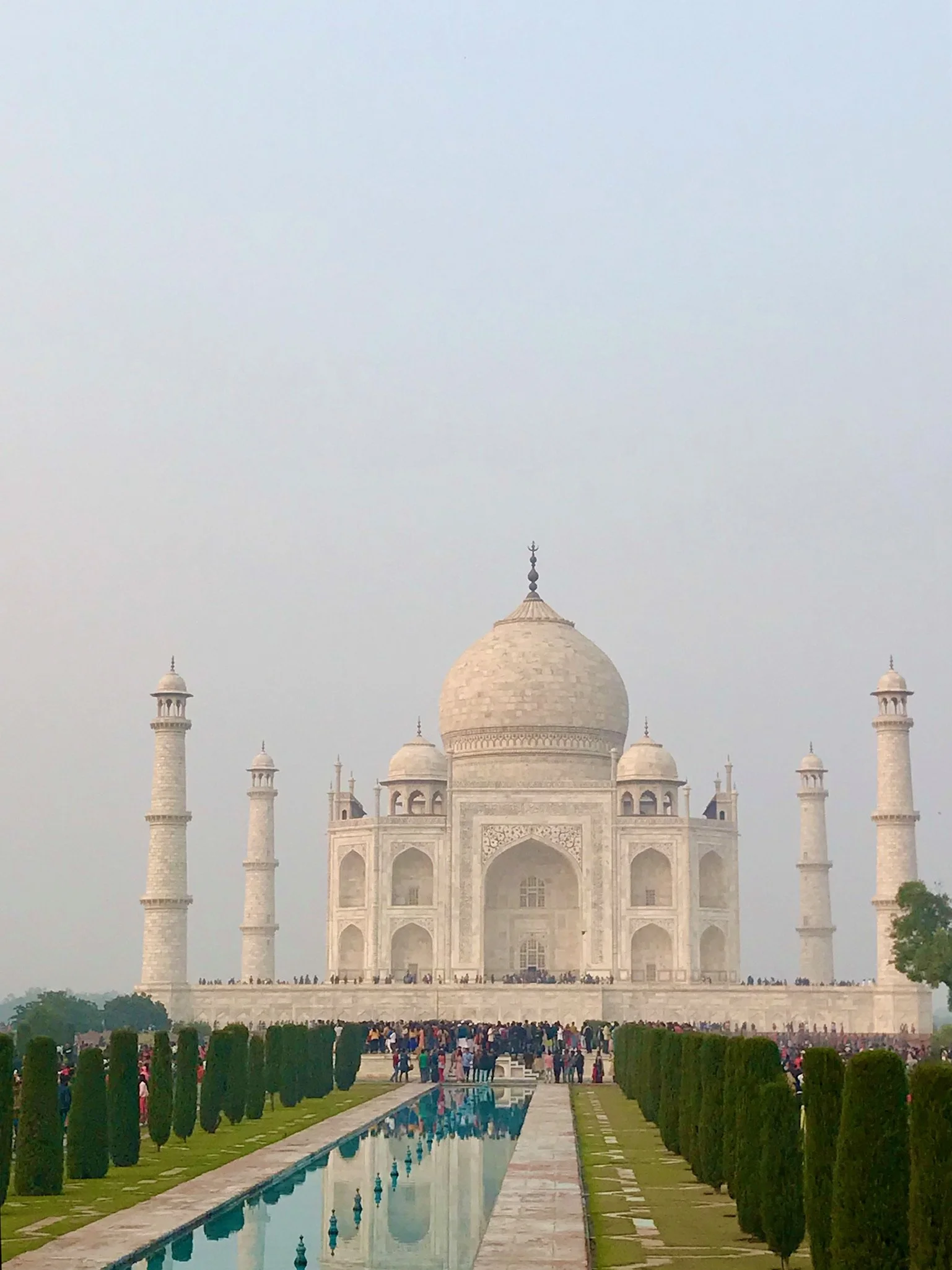 The Taj Mahal, a white marble mausoleum with a large central dome, surrounded by four minarets, on a cloudy day with a large foreground reflecting pool and a crowd of visitors.