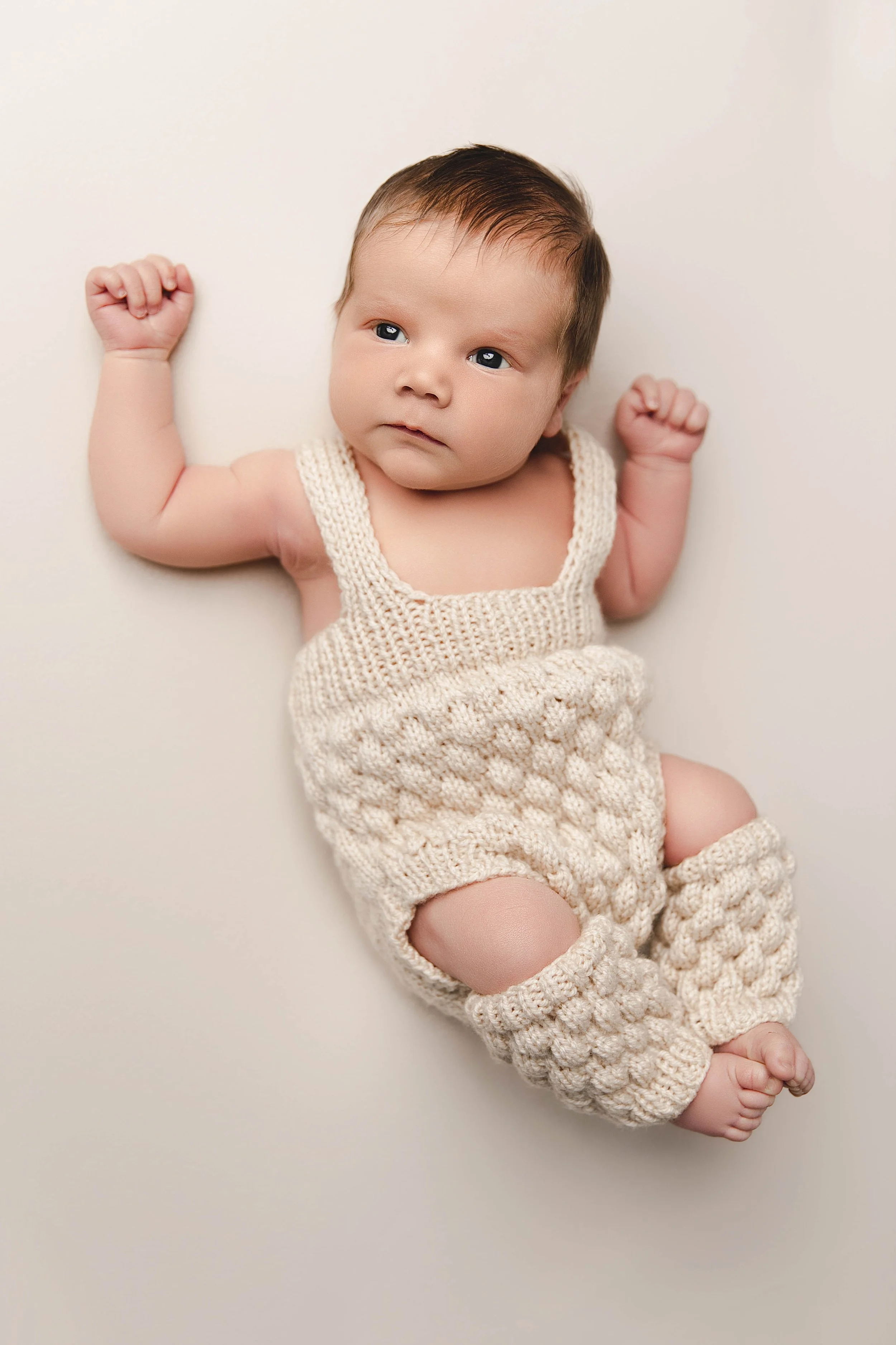 A baby lying down on a white surface, wearing a cream-colored knitted outfit, with fists raised and a neutral expression.