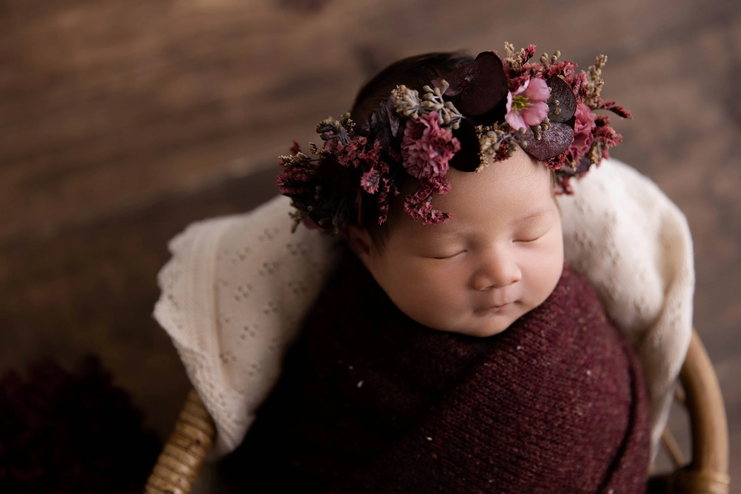 A sleeping baby wearing a dark burgundy knitted outfit and a floral crown with dark purple and pink flowers, resting in a wicker chair with a white textured blanket backrest, on a wooden floor.