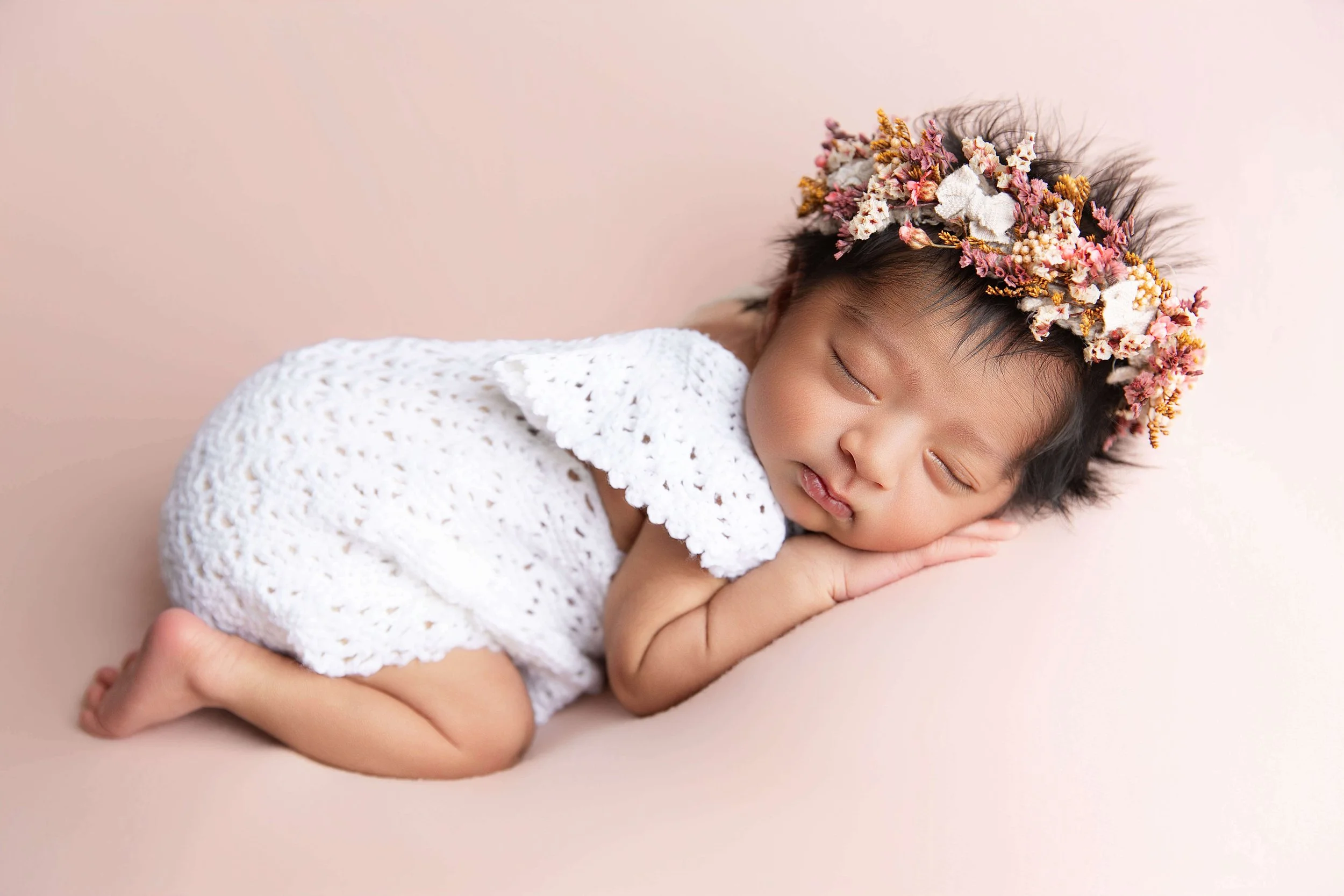 A peacefully sleeping baby girl with a floral crown, lying on her stomach on a soft pink surface, wearing a white lace dress.