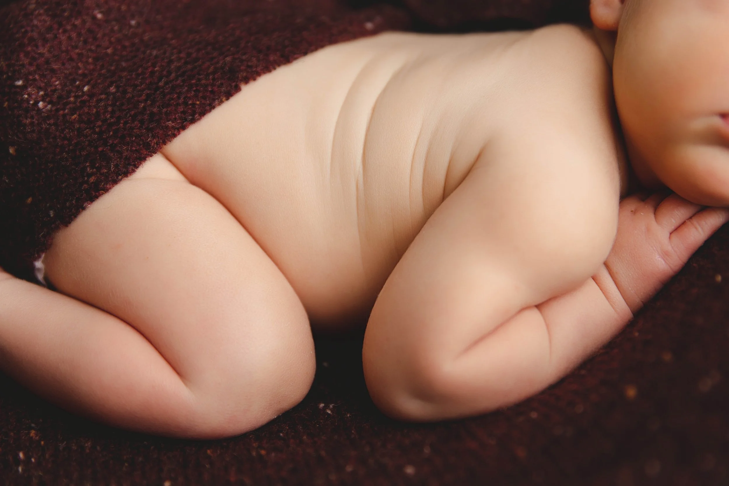Close-up of a baby lying on a brown textured blanket, showing the baby's smooth chubby arm, hand, and face.