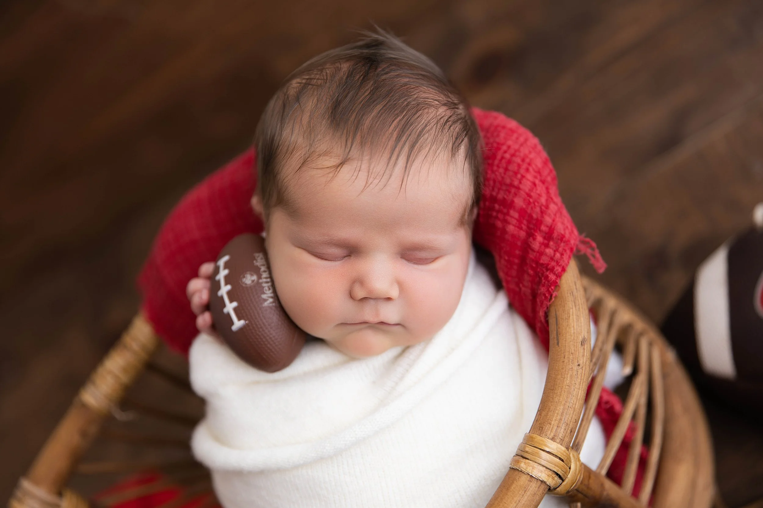 A sleeping baby with brown hair, wrapped in a white blanket, resting on a small basket with a red blanket. The baby is holding a small football-shaped object.
