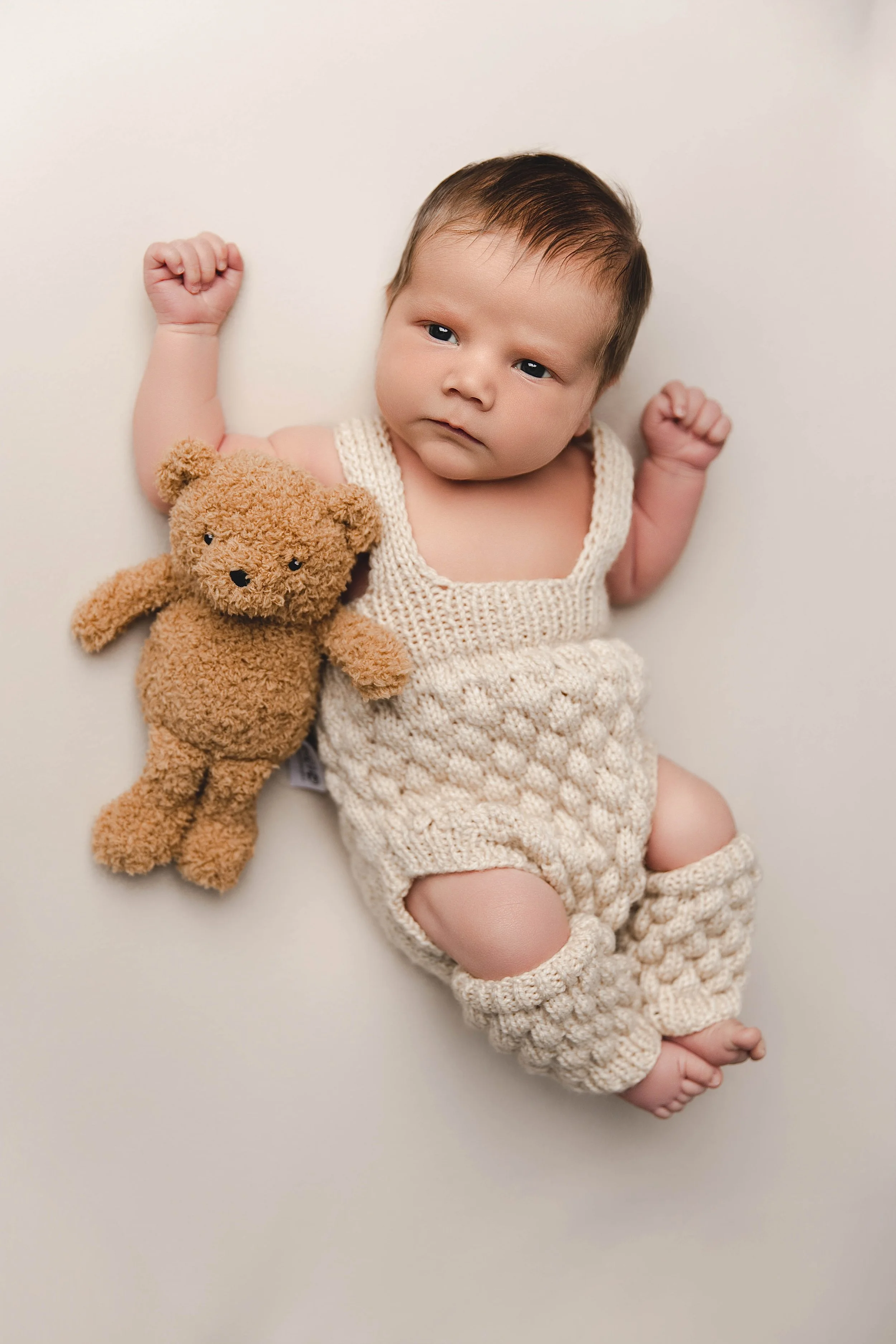 A baby lying on a white surface holding a brown teddy bear, wearing a cream-colored knitted outfit, with fists raised and looking at the camera.