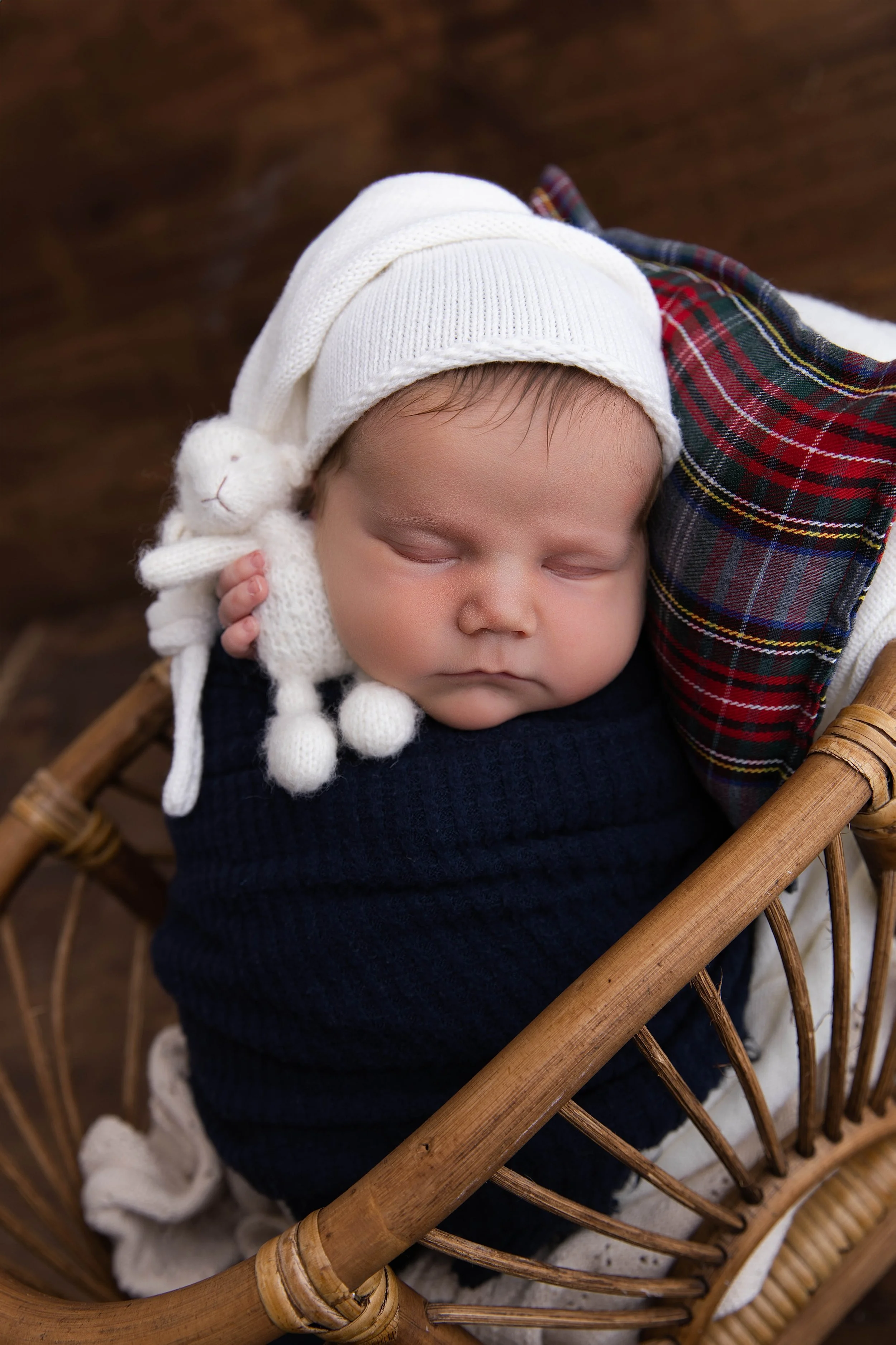 A sleeping baby wearing a white knit hat with a bunny ear and pom-pom details, wrapped in dark blue cloth, resting in a wicker basket with a plaid blanket nearby.