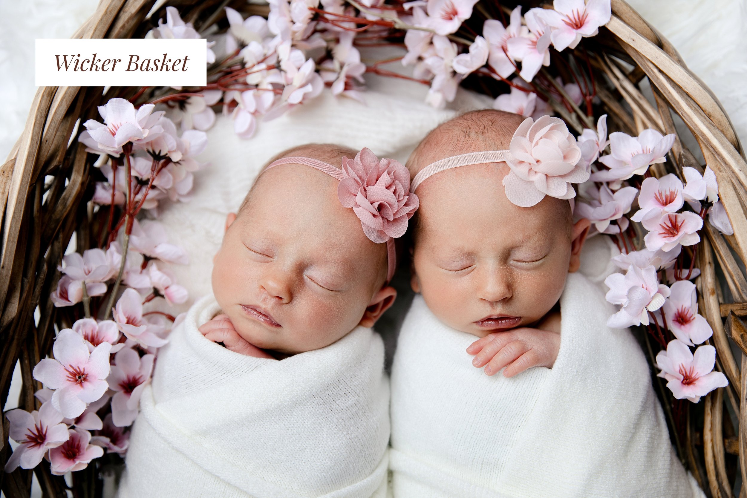 Two newborn babies swaddled in white blankets, sleeping in a woven basket surrounded by pink flowers and wearing pink headbands with fabric flowers.