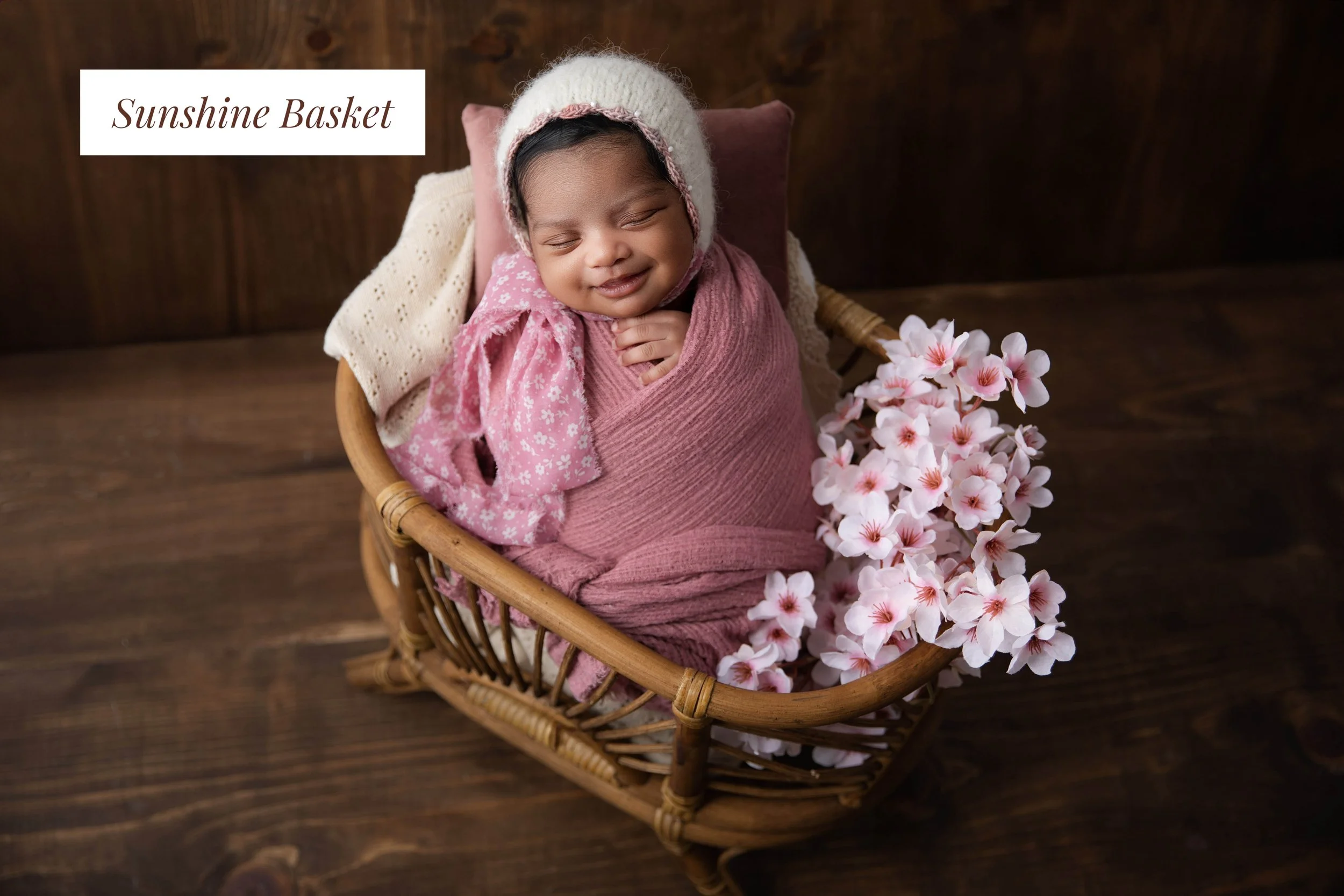 A smiling baby wrapped in pink cloth, wearing a white knit hat, sitting in a wicker basket surrounded by pink flowers, with a wooden background.