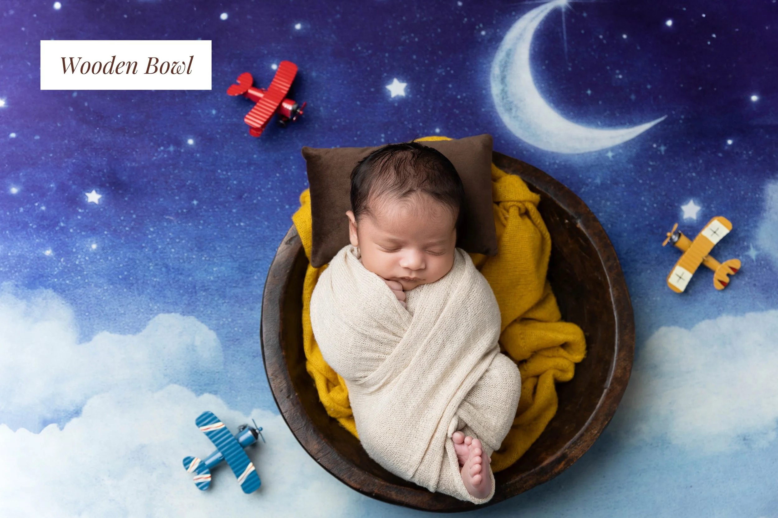A sleeping baby wrapped in a beige blanket, lying in a wooden bowl on a blue starry sky background with a moon and clouds design, surrounded by small toy airplanes and a red toy airplane.