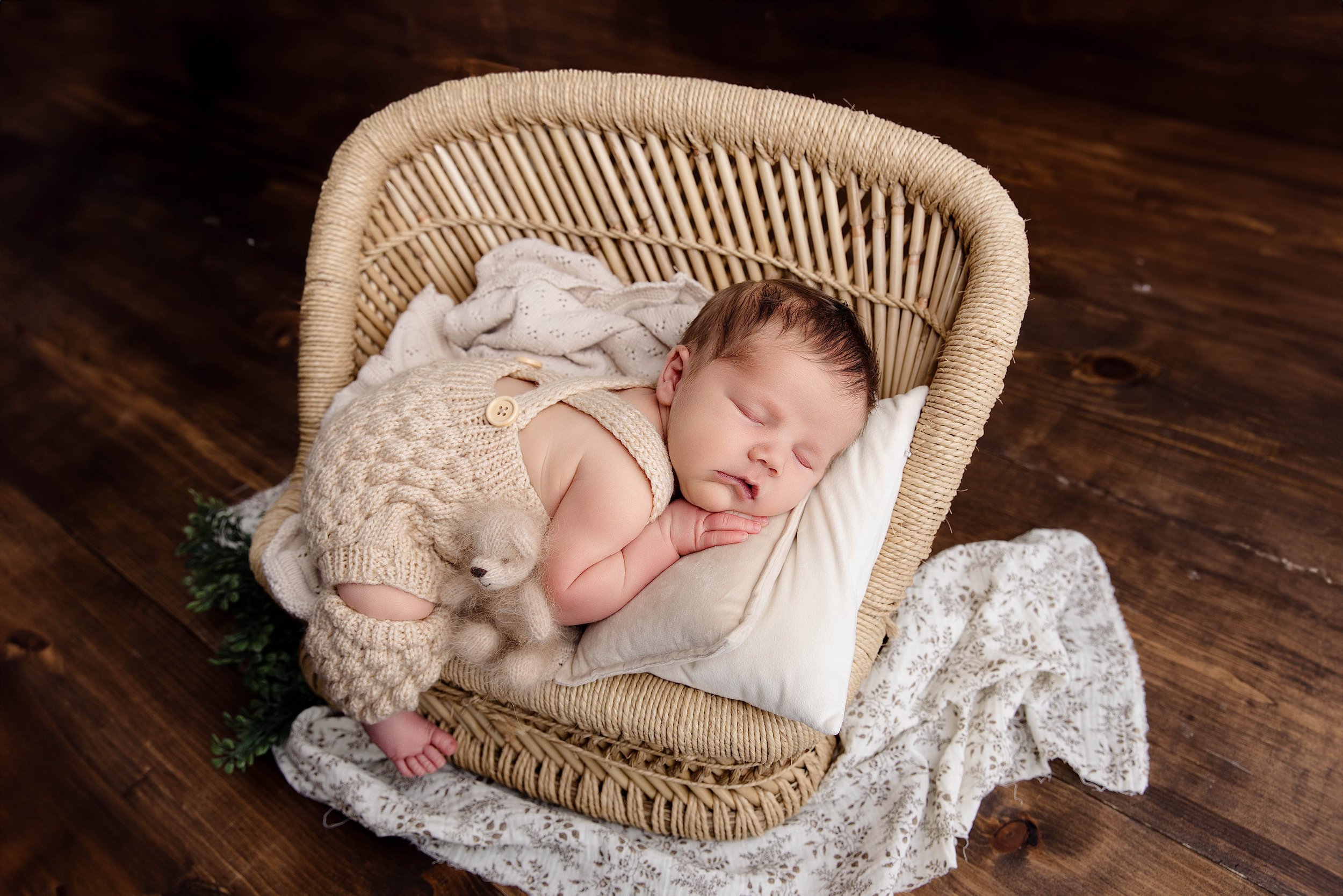 A sleeping newborn baby lying on a small pillow in a wicker bassinet with a knitted outfit and tiny stuffed bear, on a wooden floor with a lace cloth underneath.